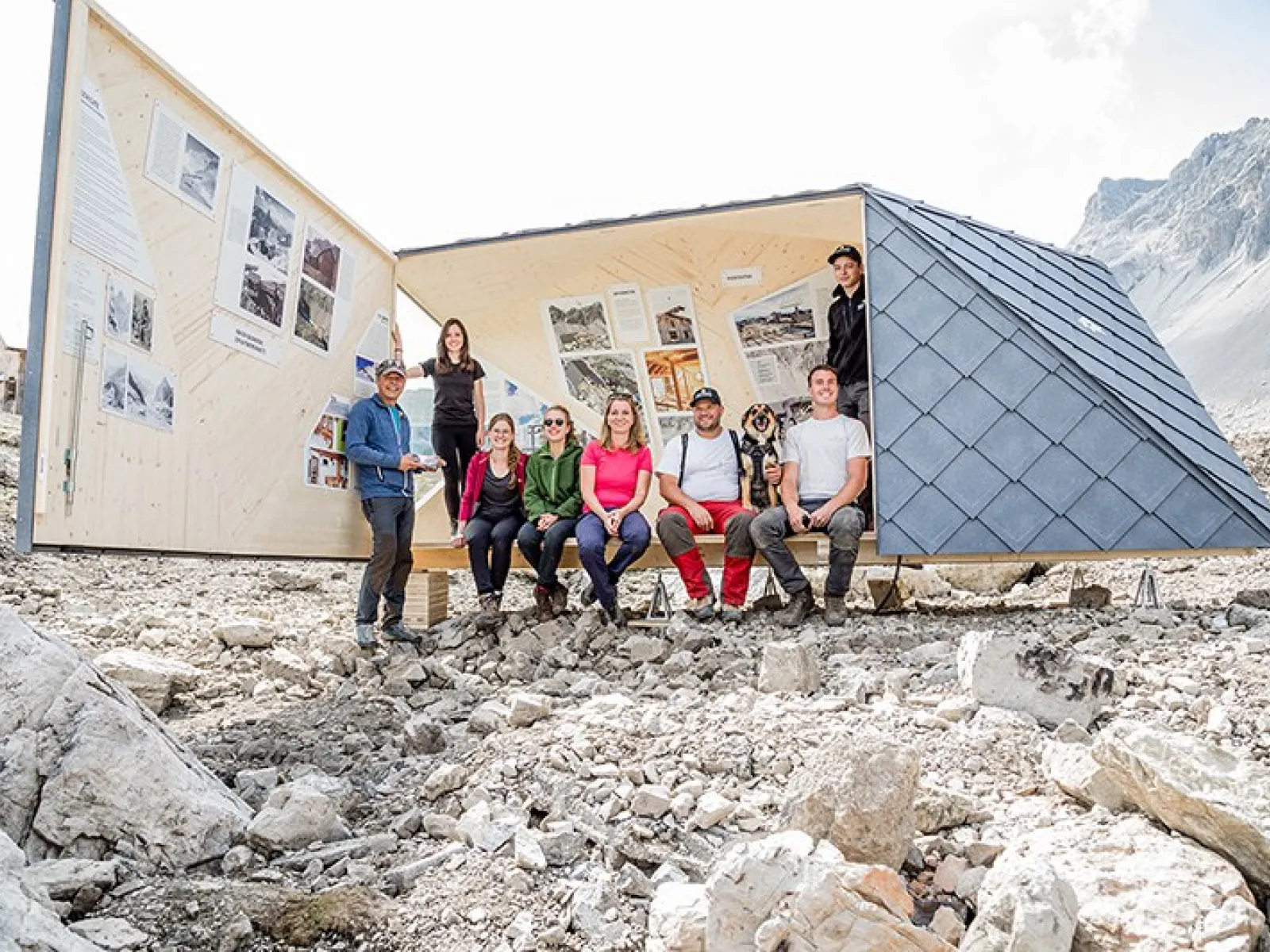 Eine Gruppe von Studierenden sitzt vor einer modernen, dreieckigen Bergunterkunft, die als architektonisches Ausstellungsprojekt dient und nachhaltige Baupraktiken in einer felsigen Berglandschaft zeigt.