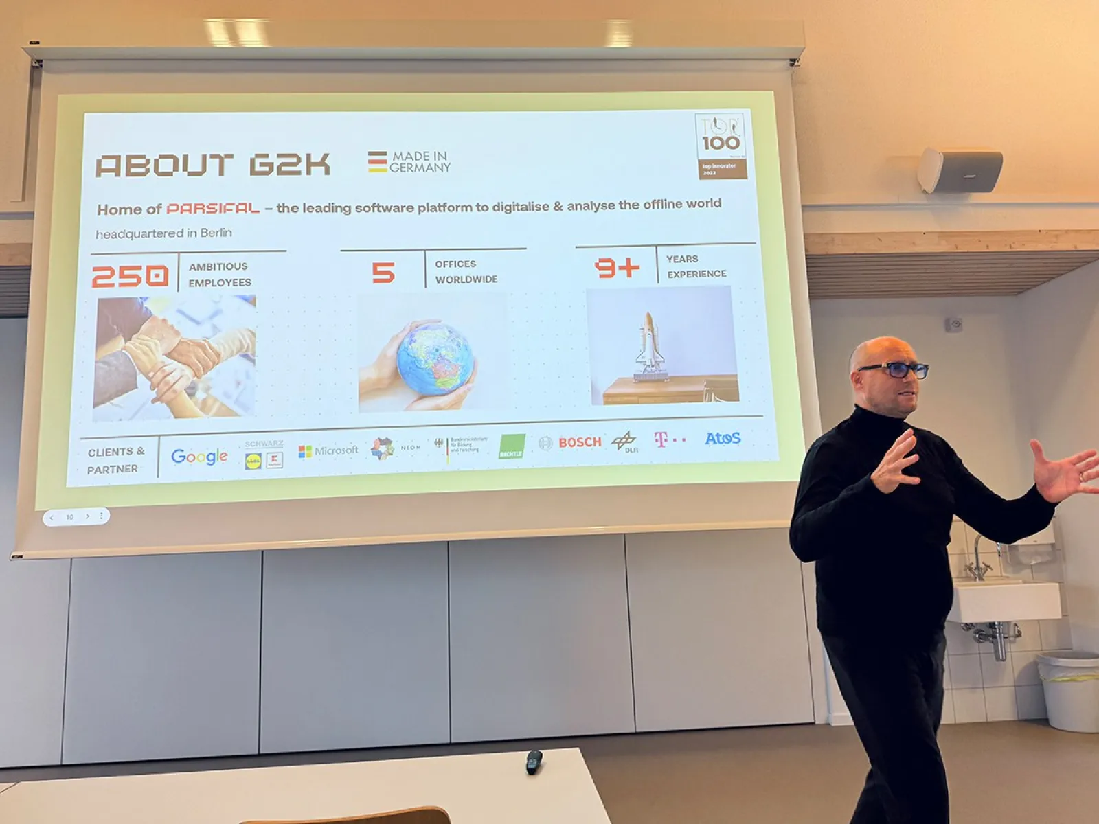 Ein Mann hält einen Vortrag vor einer Präsentationsfolie. Die Folie zeigt Informationen über das Unternehmen G2K, darunter die Anzahl der Mitarbeiter, Standorte und Jahre Erfahrung. Logos von Partnerunternehmen sind ebenfalls sichtbar.