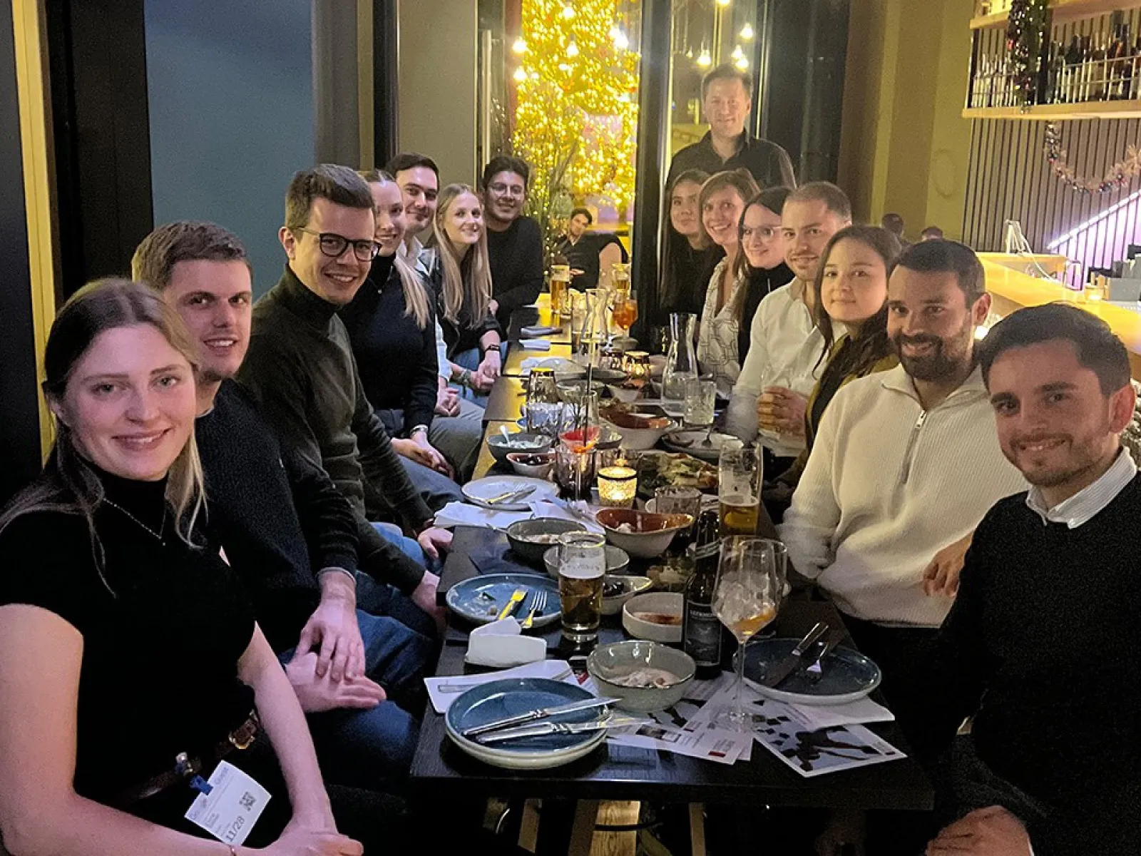 Eine Gruppe von Alumni der Universität Liechtenstein sitzt in einem Restaurant an einem langen Tisch, lächelnd und in geselliger Runde. Auf dem Tisch stehen Getränke und Teller mit Speisen. Die Atmosphäre ist warm und einladend.