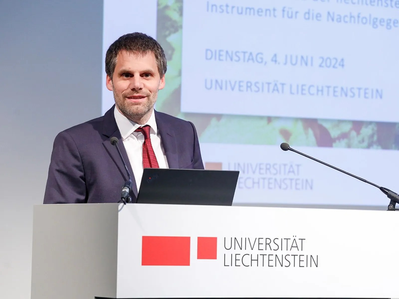 Ein Mann in einem Anzug steht an einem Rednerpult mit dem Logo der Universität Liechtenstein. Im Hintergrund ist eine Präsentationsfolie mit dem Datum 4. Juni 2024 zu sehen.