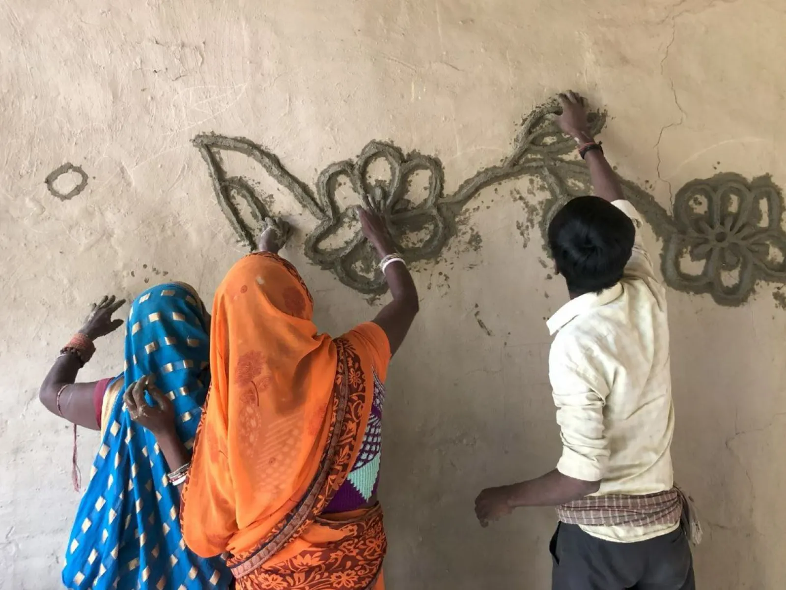 Drei Personen gestalten gemeinsam eine Wand mit traditionellen Mustern aus Lehm. Sie tragen bunte Kleidung und arbeiten konzentriert an einem floralen Design.