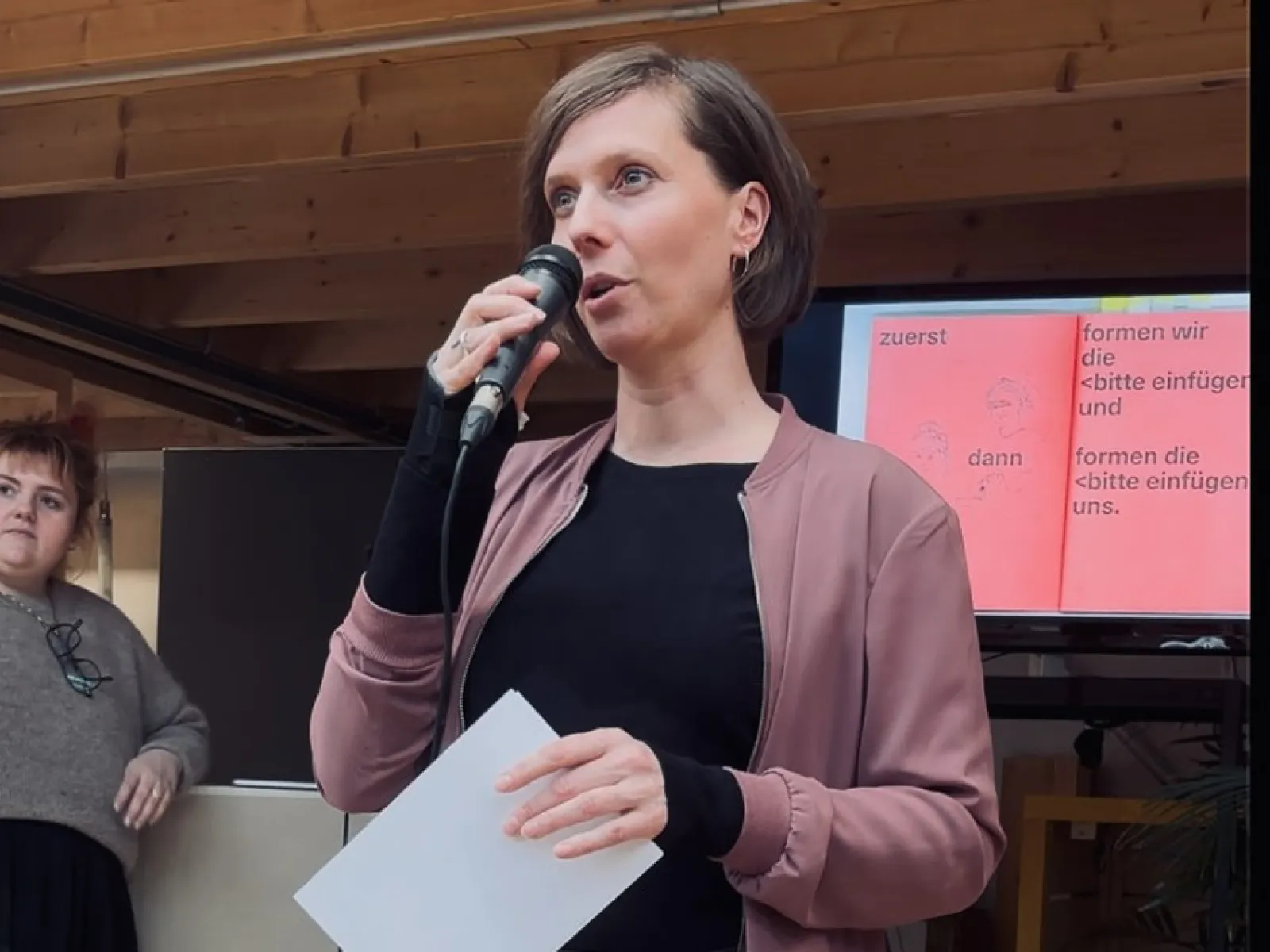 Eine Person hält ein Mikrofon und spricht vor einem Publikum. Sie trägt eine rosa Jacke und hält ein Blatt Papier. Im Hintergrund ist eine Präsentation auf einem Bildschirm zu sehen. Eine weitere Person steht links daneben und hört zu.