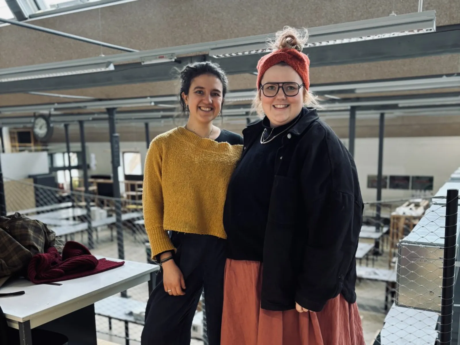 Zwei Personen stehen lächelnd in einem modernen Architekturatelier mit großen Fenstern und industriellem Design. Sie tragen lässige, kreative Kleidung. Im Hintergrund sind Arbeitsplätze und eine Uhr sichtbar.