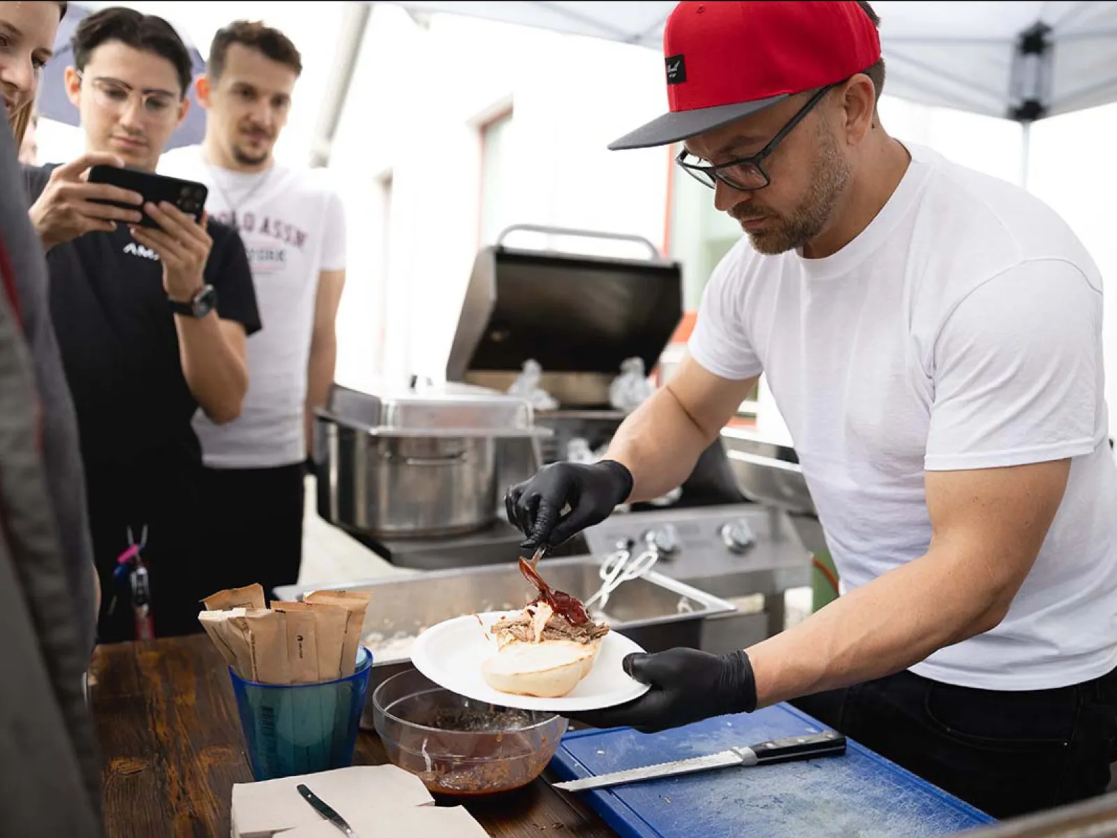 Ein Mann mit roter Kappe und schwarzen Handschuhen bereitet Essen auf einem Teller vor. Im Hintergrund stehen mehrere Personen und beobachten ihn. Eine Grillstation ist ebenfalls zu sehen. Die Szene findet im Freien unter einem Zelt statt.
