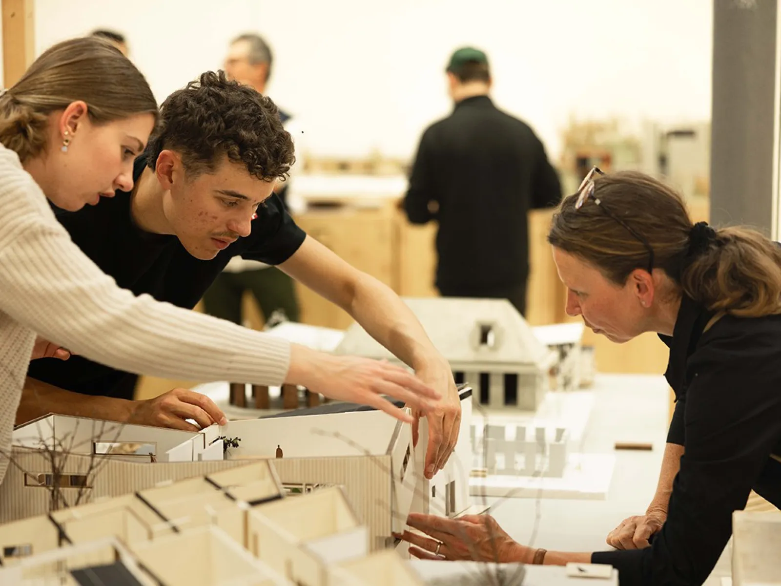 Drei Personen betrachten und diskutieren über Architekturmodelle auf einem Tisch in einem Ausstellungsraum. Im Hintergrund sind weitere Personen und Modelle zu sehen.