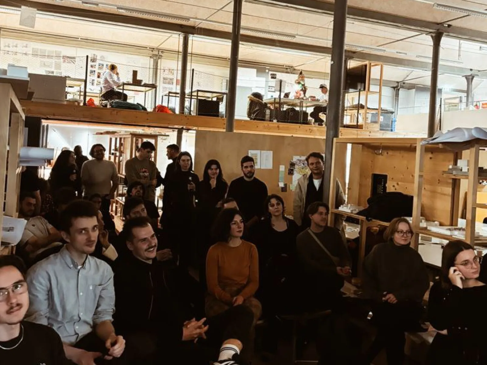 Eine Gruppe von Studierenden und Alumni sitzt in einem Architekturstudio und hört einer Präsentation zu. Im Hintergrund sind Arbeitsplätze und eine Uhr an der Wand zu sehen. Die Atmosphäre ist konzentriert und interessiert.