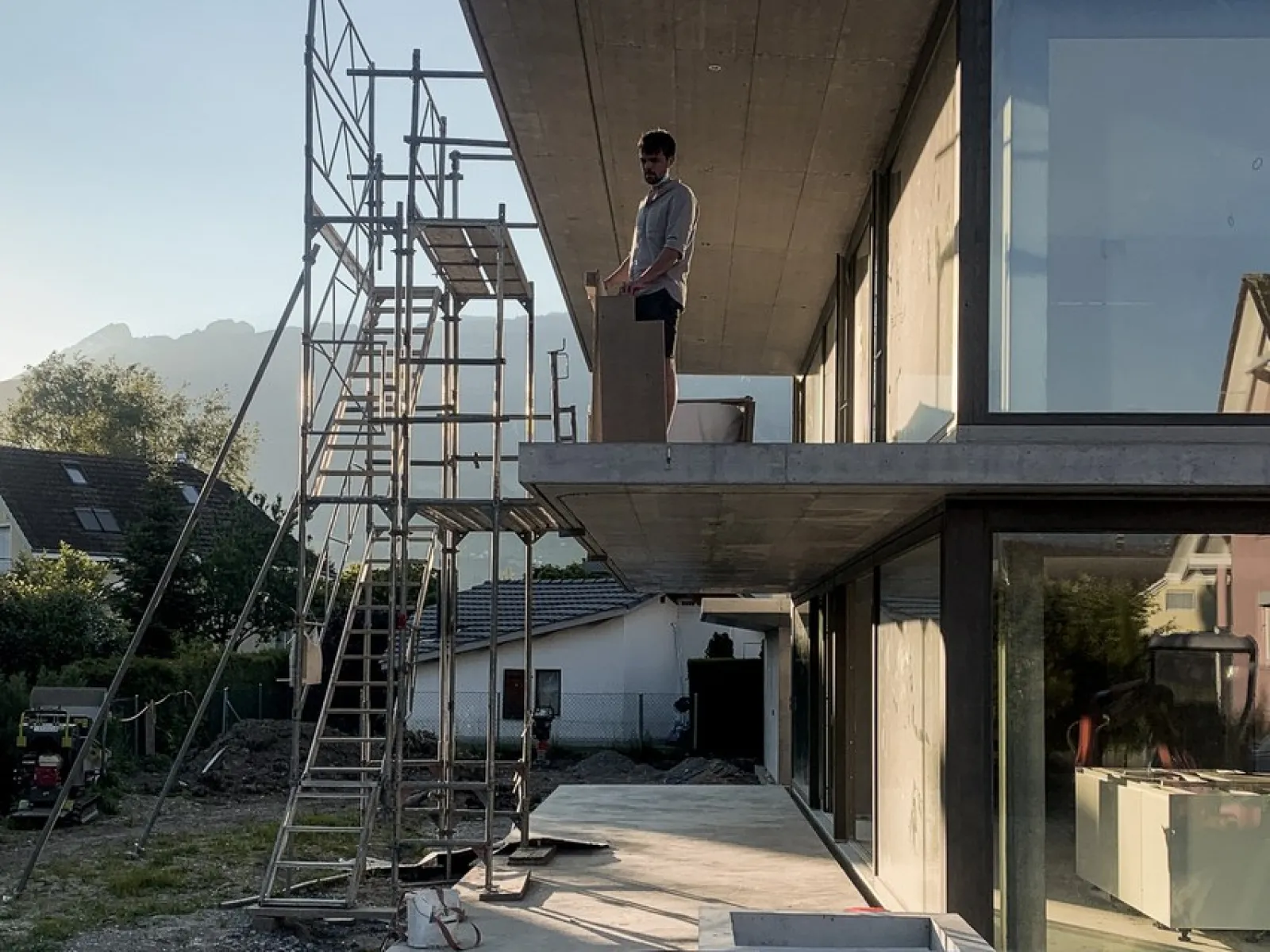 Ein Architekt steht auf einem modernen Gebäude mit großen Glasfenstern, daneben ein Baugerüst. Im Hintergrund sind Berge und ein Haus zu sehen. Die Szene zeigt eine Baustelle bei Sonnenuntergang.