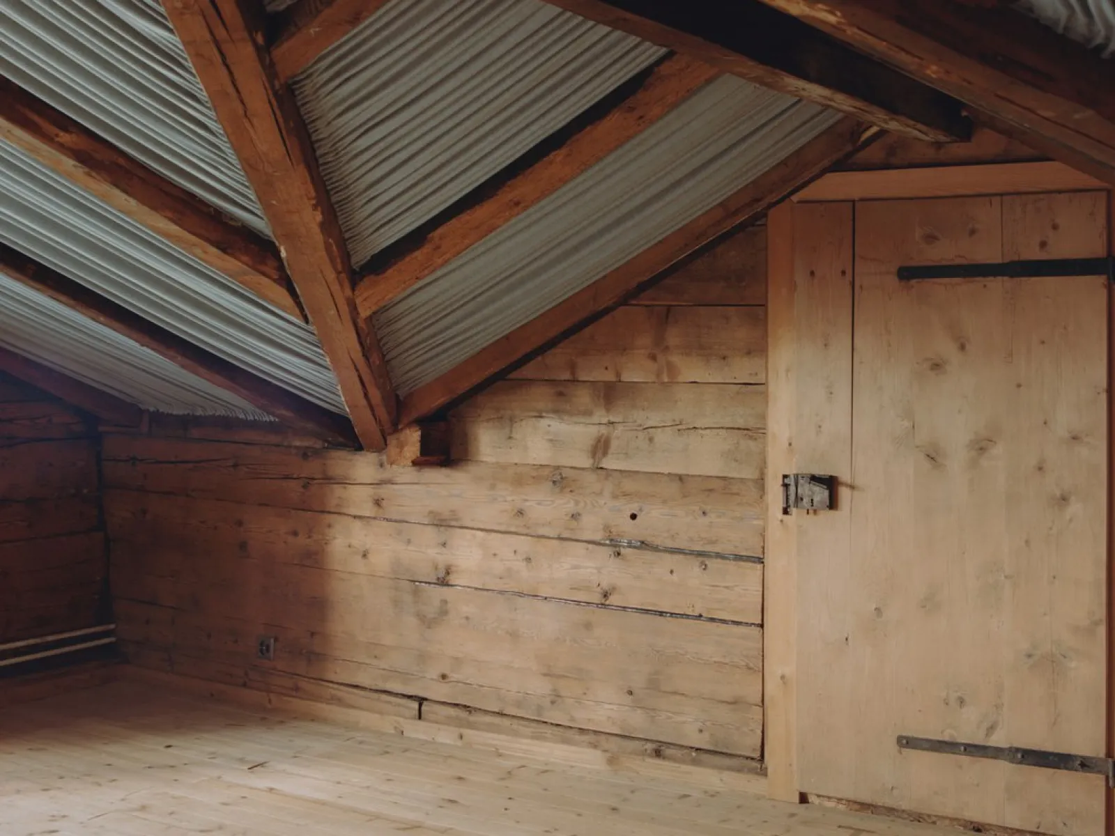 Innenansicht eines Dachbodens mit Holzwänden und Holzboden, sichtbare Dachbalken und Wellblechverkleidung. Eine Holztür befindet sich an der rechten Wand.