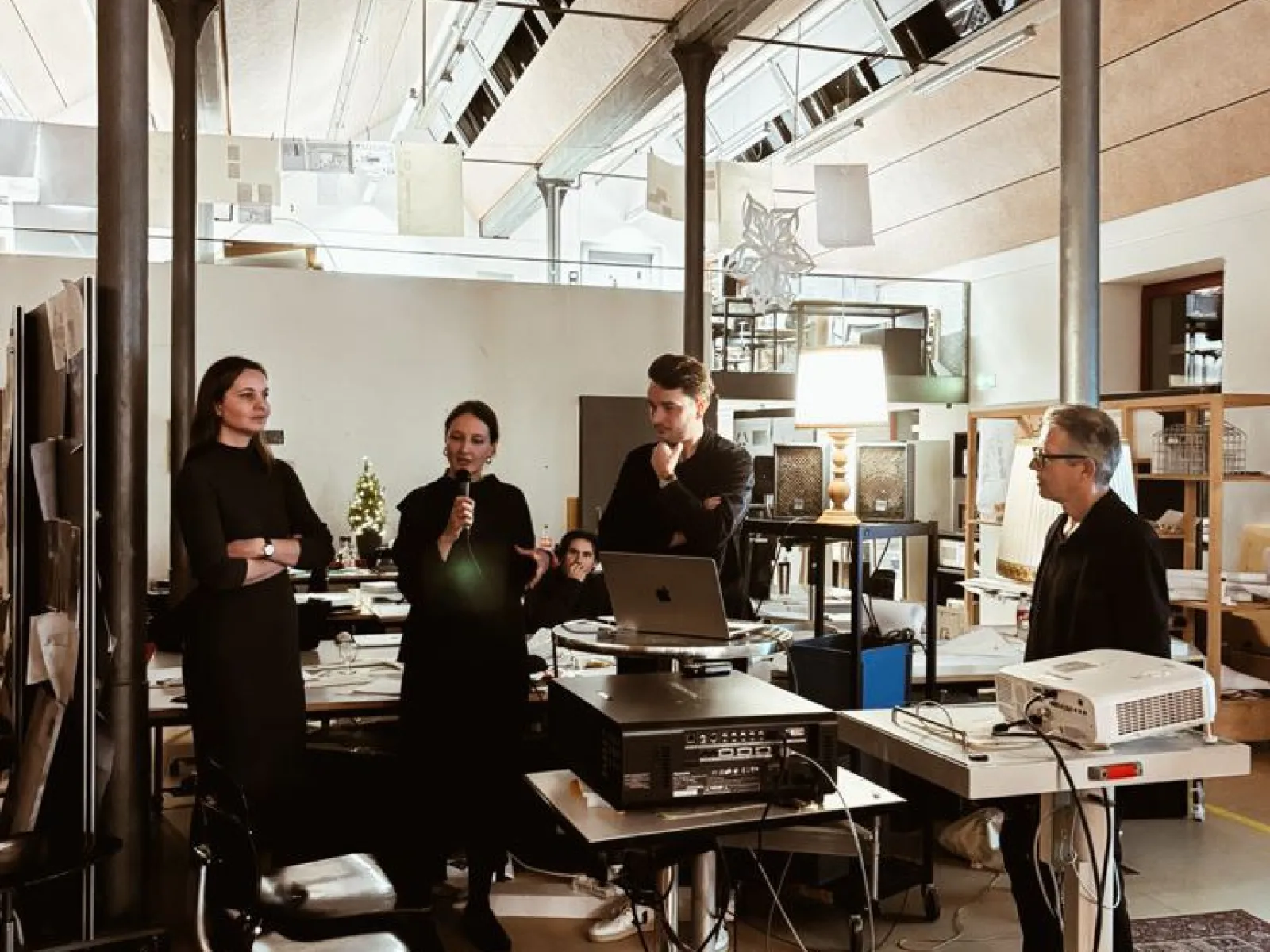 Eine Gruppe von Personen steht in einem modernen Architekturstudio. Eine Person spricht in ein Mikrofon, während andere aufmerksam zuhören. Im Raum sind technische Geräte und Präsentationsmaterialien zu sehen.