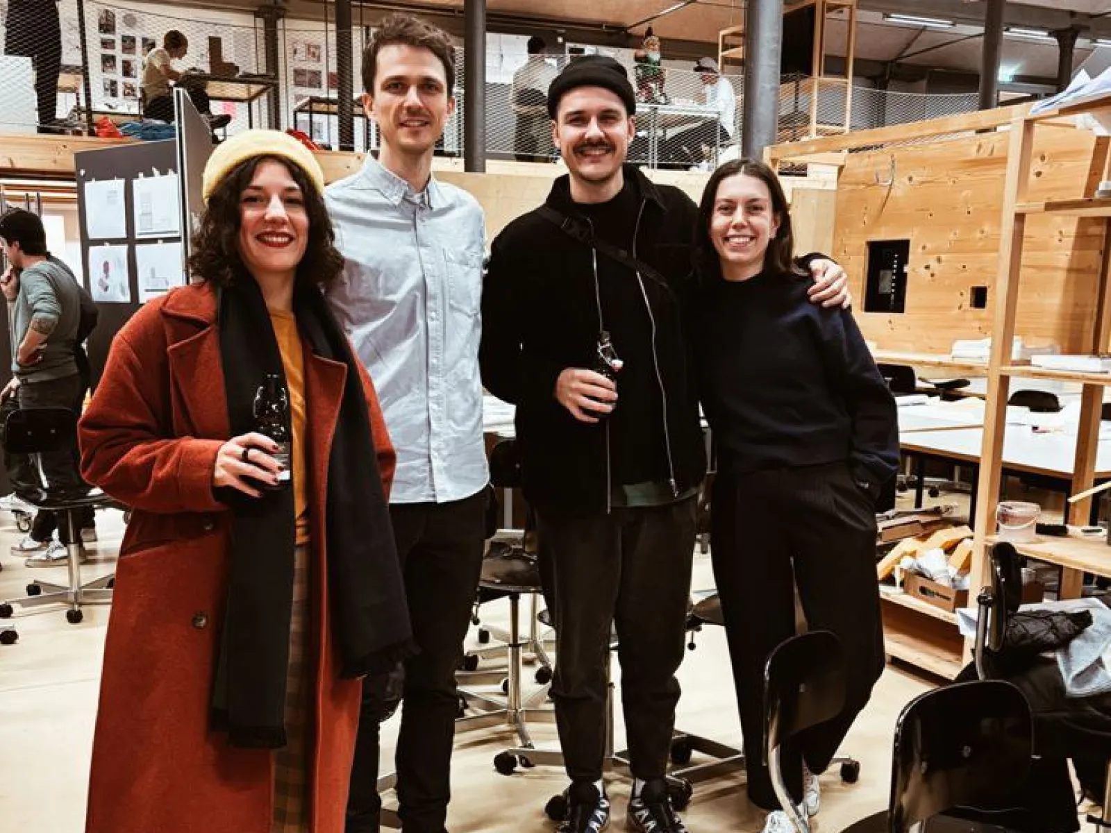 Vier Personen stehen lächelnd in einem Architekturstudio. Sie tragen lässige Kleidung und halten Getränke in der Hand. Im Hintergrund sind Arbeitsplätze und Modelle zu sehen. Die Atmosphäre wirkt freundlich und kreativ.