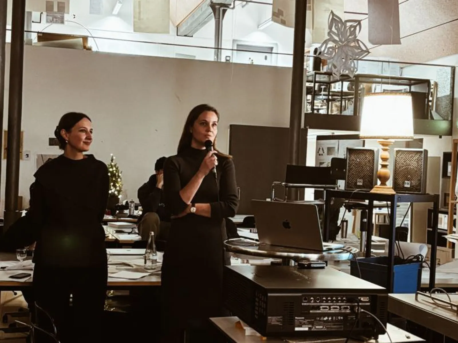 Zwei Personen stehen in einem modernen Architekturstudio. Eine Person hält ein Mikrofon und spricht, während die andere zuhört. Im Hintergrund sind Arbeitsplätze und Architekturmodelle zu sehen.