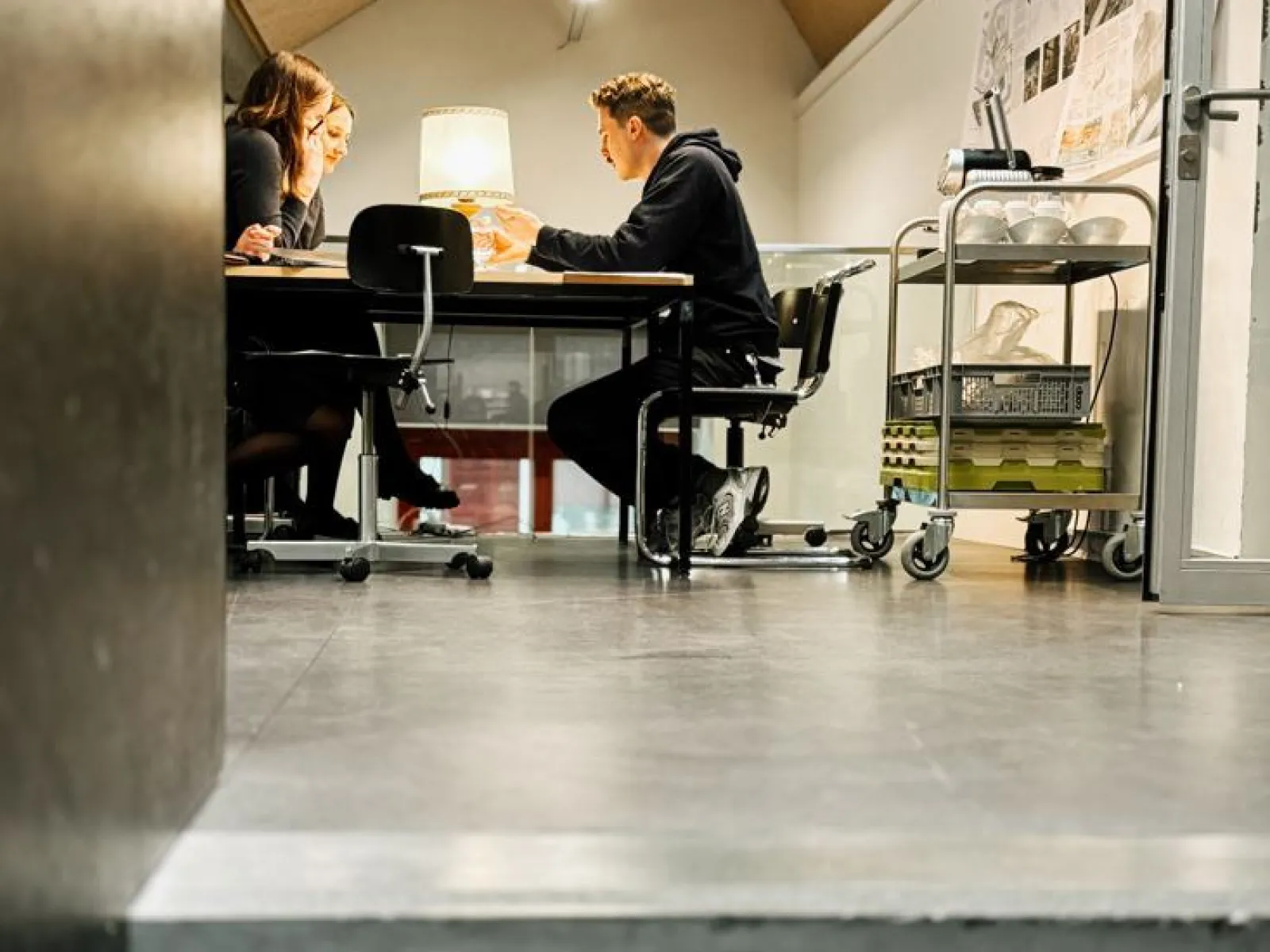 Drei Personen sitzen an einem Tisch in einem modernen Seminarraum und diskutieren. Im Hintergrund sind Architekturpläne an der Wand sichtbar. Der Raum ist hell beleuchtet und wirkt einladend.