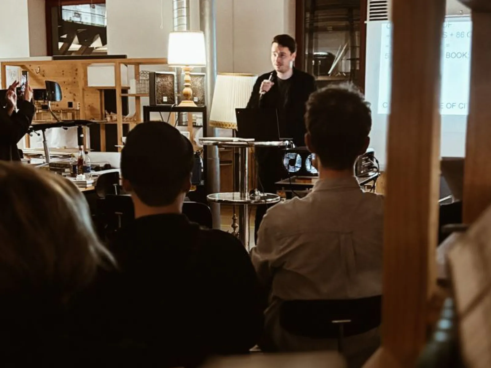Ein Vortragender spricht vor einer Gruppe von Studierenden in einem Raum mit Präsentationstechnik. Im Hintergrund sind Regale und eine Lampe sichtbar. Die Atmosphäre wirkt konzentriert und lernorientiert.