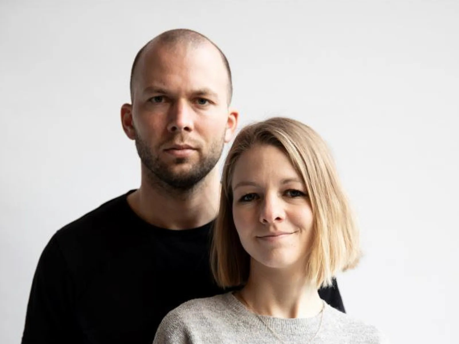 Ein Mann und eine Frau stehen vor einem neutralen Hintergrund. Beide blicken direkt in die Kamera. Die Frau trägt einen grauen Pullover, der Mann ein schwarzes T-Shirt.