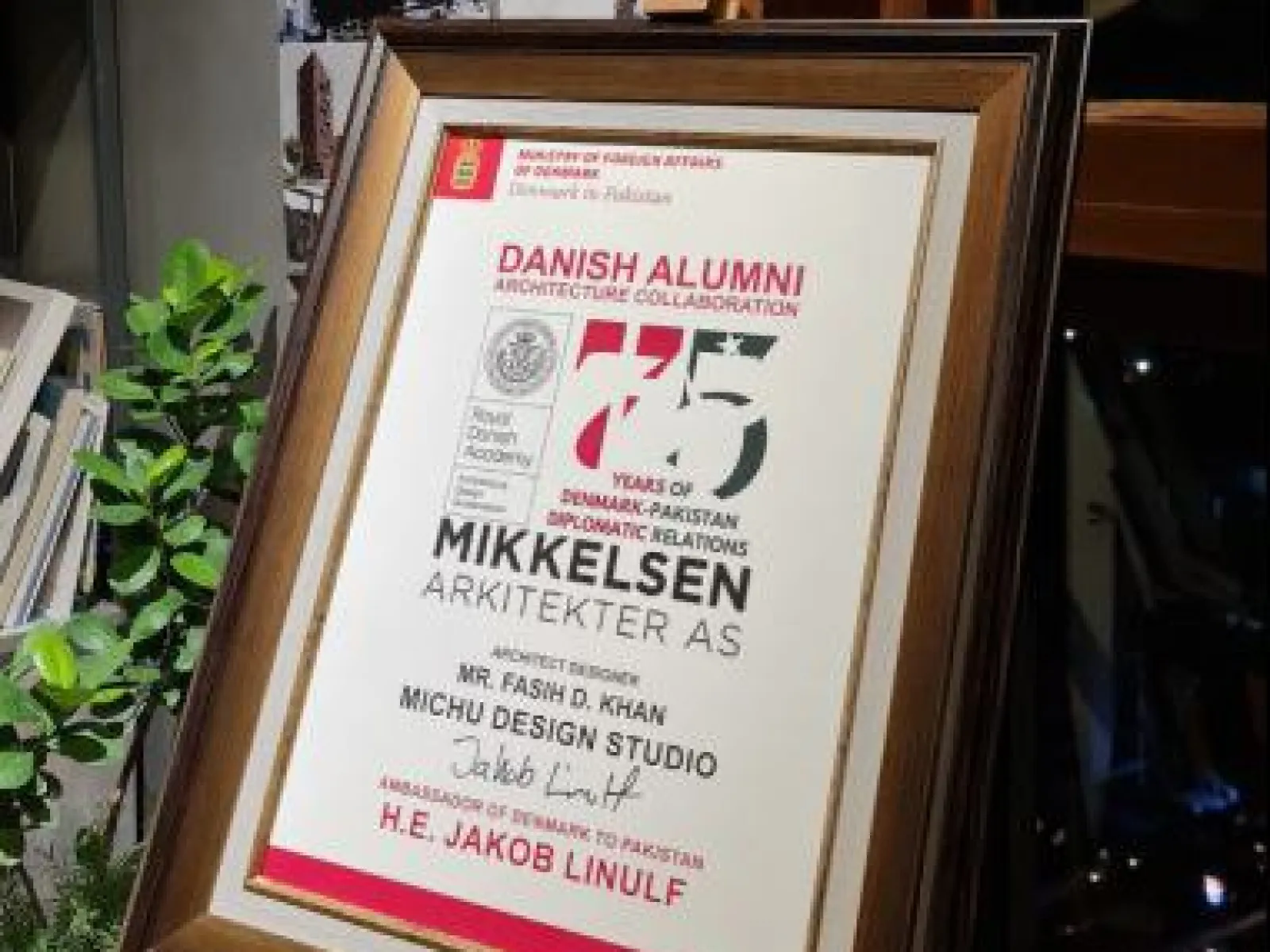 Ein gerahmtes Zertifikat zur Anerkennung der Zusammenarbeit im Bereich Architektur zwischen Dänemark und Pakistan, ausgestellt an Fasih D. Khan, steht auf einem Regal. Im Hintergrund sind Architekturprojekte und Gruppenfotos zu sehen.