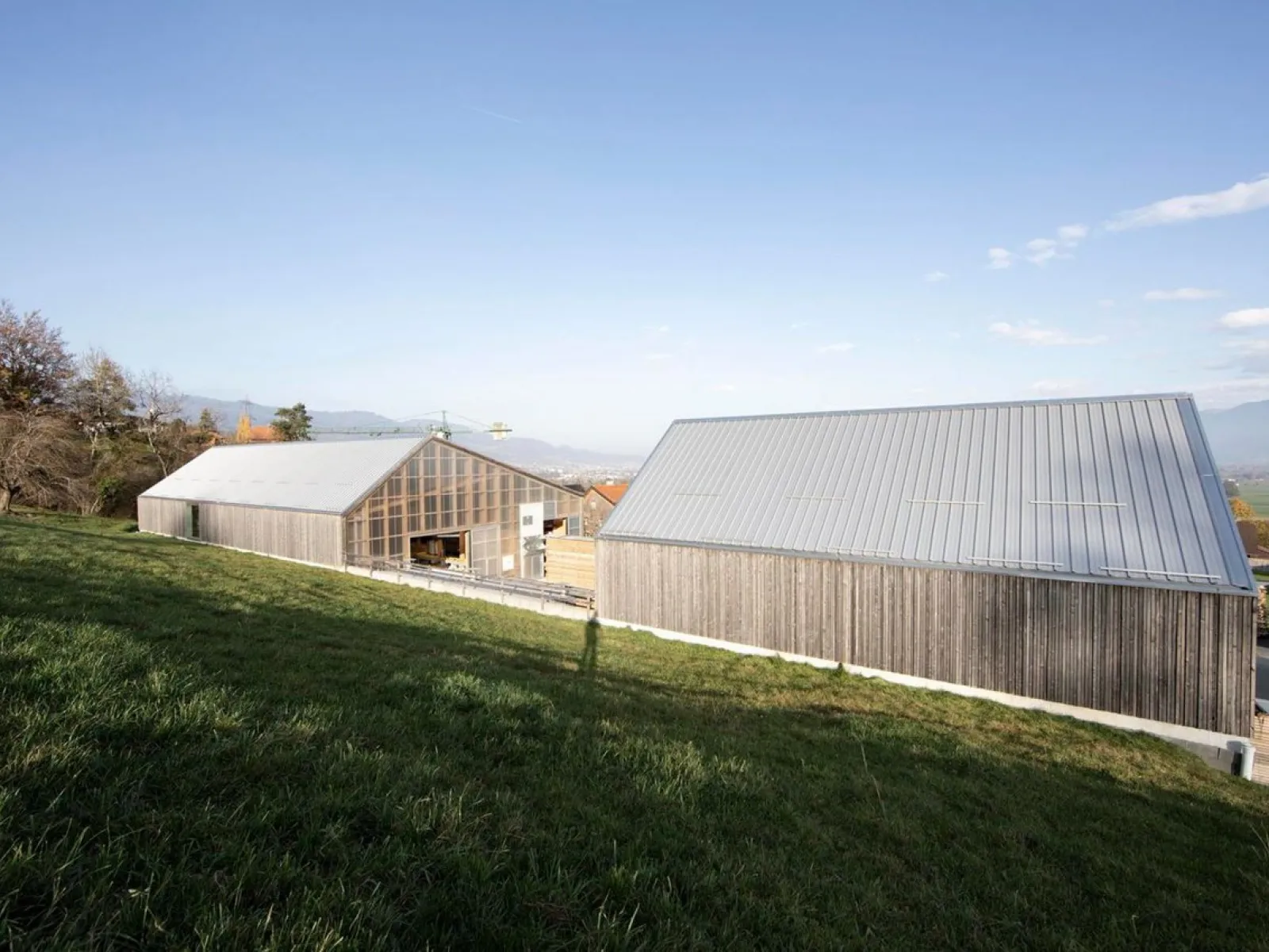 Zwei moderne Gebäude mit Holz- und Glasfassaden in einer ländlichen Umgebung. Die Architektur ist schlicht und kontextbezogen, eingebettet in eine grüne Landschaft.