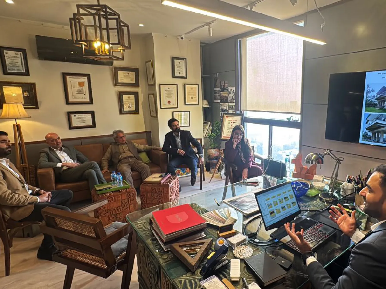 Ein Büro mit mehreren Personen, die in einem Meeting sitzen. Eine Person sitzt an einem Schreibtisch mit einem Laptop, während andere auf Stühlen und einem Sofa Platz genommen haben. An der Wand hängen Zertifikate und ein Bildschirm zeigt architektonische Entwürfe.