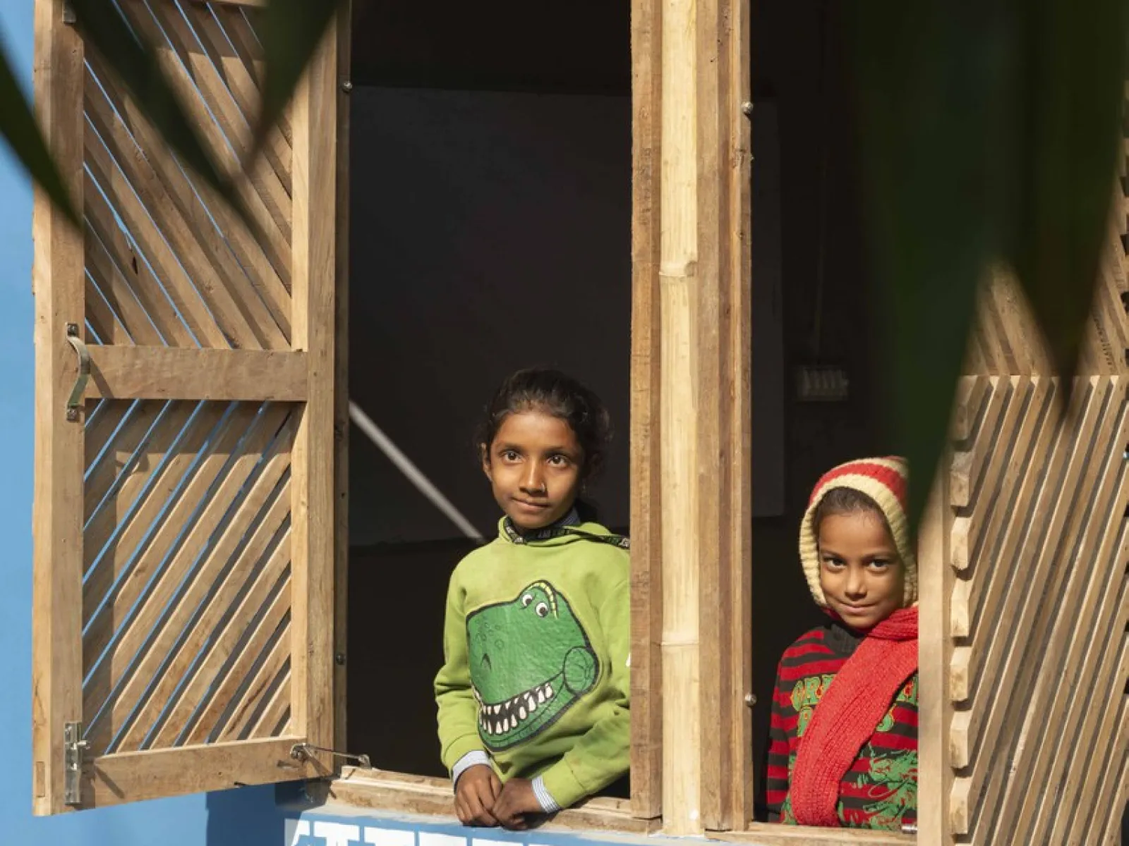 Zwei Kinder schauen aus einem offenen Fenster eines Gebäudes mit blauer Wand und dekorativem Muster. Das Fenster hat hölzerne Fensterläden. Im Vordergrund sind unscharfe Blätter zu sehen.
