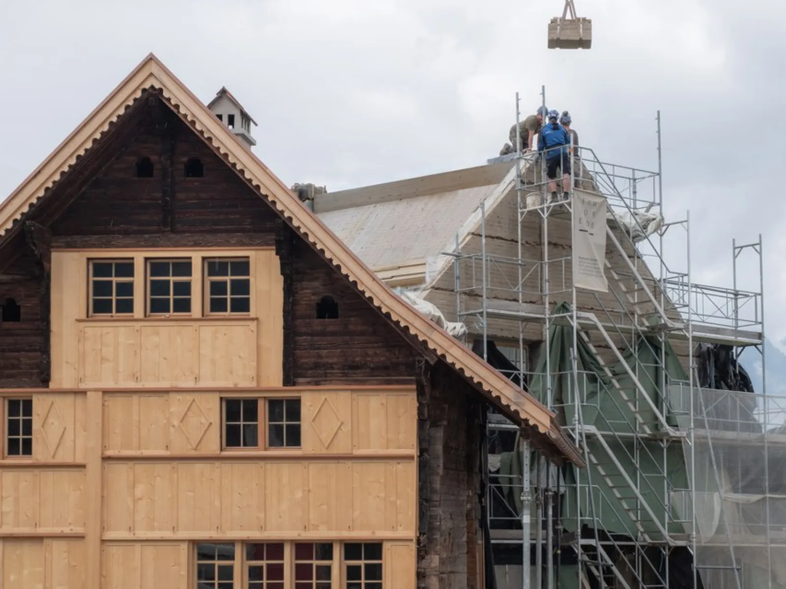 Ein traditionelles Gebäude wird renoviert. Links ist eine Holzfassade zu sehen, rechts stehen Gerüste mit Bauarbeitern auf dem Dach. Der Himmel ist bewölkt.