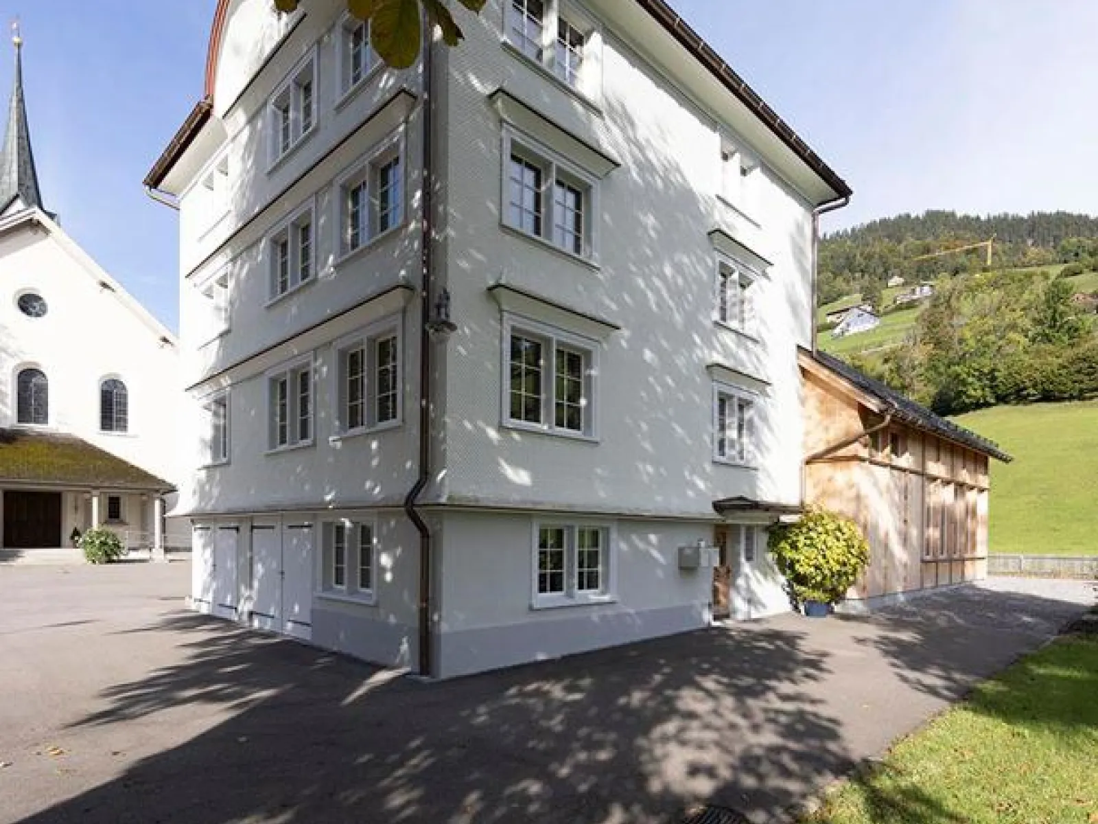 Ein weißes, mehrstöckiges Gebäude mit traditionellen Fenstern steht neben einer Kirche im ländlichen St. Galler Rheintal. Im Hintergrund sind grüne Hügel zu sehen. Der Schatten eines Baumes fällt auf den gepflasterten Vorplatz.