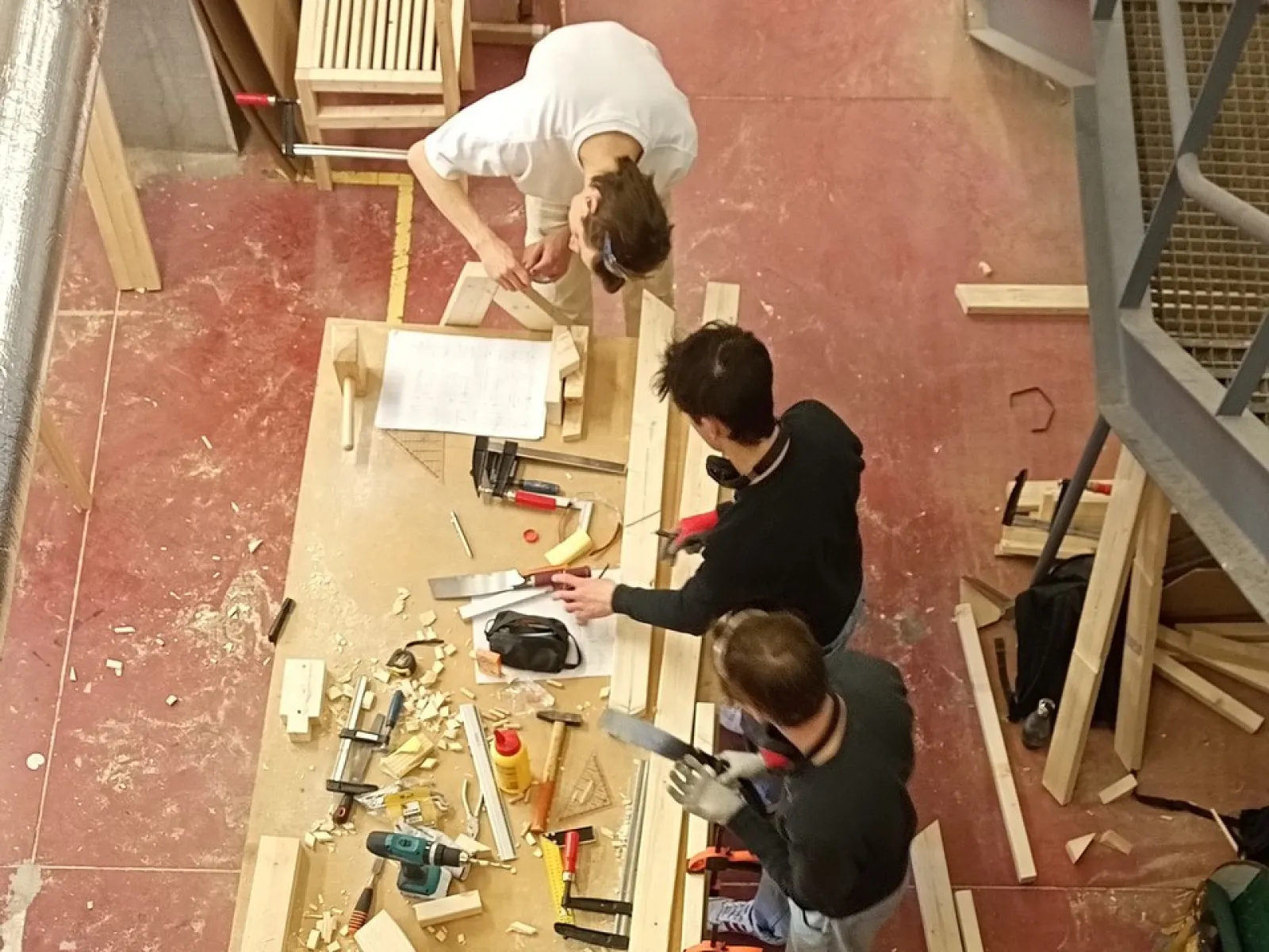 Vier Studierende arbeiten gemeinsam in einer Werkstatt an einem Holztisch. Sie sind von oben zu sehen und verwenden verschiedene Werkzeuge, um ein Möbelstück zu bauen. Holzstücke und Werkzeuge liegen auf dem Tisch verteilt.