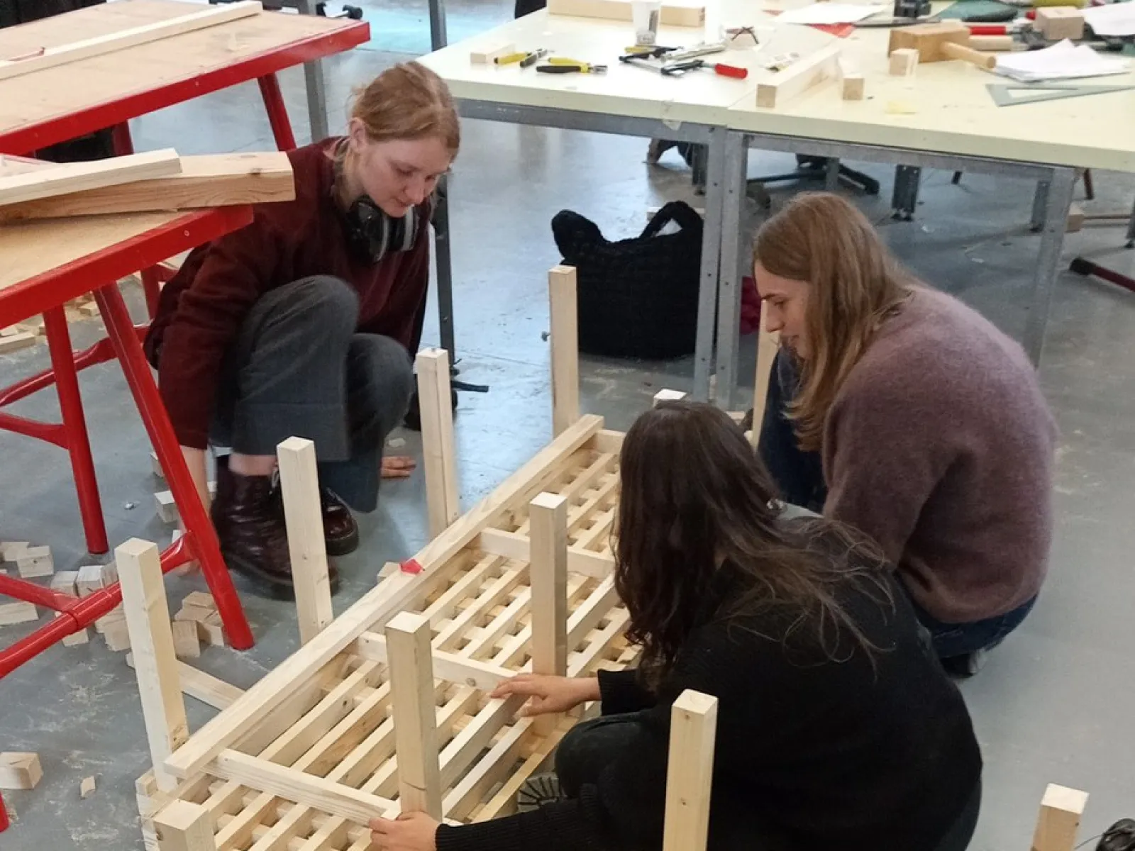Drei Studierende arbeiten gemeinsam an einem Holzprojekt in einer Werkstatt. Sie montieren ein Möbelstück aus Holzlatten. Im Hintergrund sind Werkbänke und Werkzeuge sichtbar.