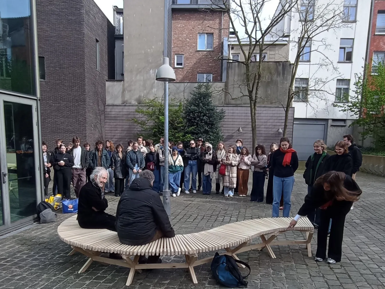Eine Gruppe von Studierenden steht im Freien um eine geschwungene Holzbank, die von zwei Personen genutzt wird. Die Szene findet auf einem gepflasterten Platz vor modernen Gebäuden statt.