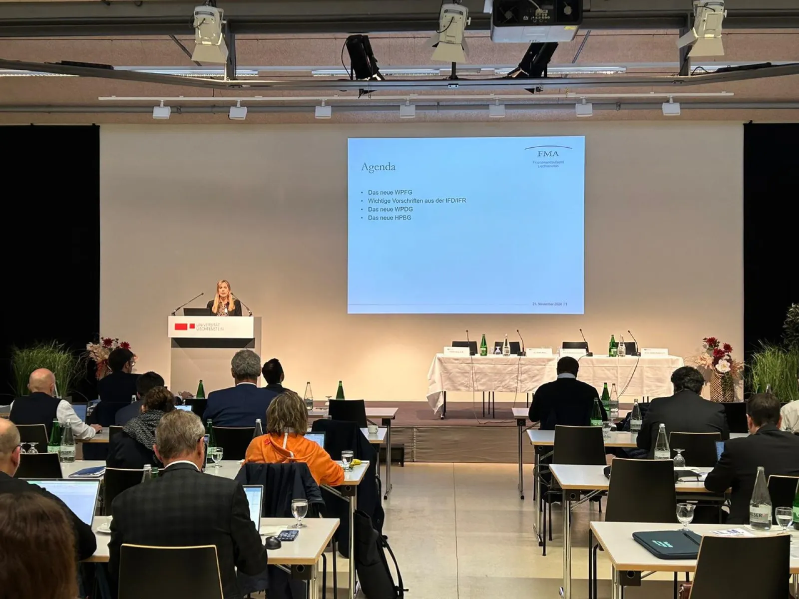 Ein Konferenzraum mit Teilnehmern, die an Tischen sitzen und einem Vortrag folgen. Eine Person steht am Rednerpult vor einer Leinwand, auf der eine Präsentation mit dem Titel "Agenda" angezeigt wird. Der Raum ist modern eingerichtet, mit mehreren Tischen und Stühlen.