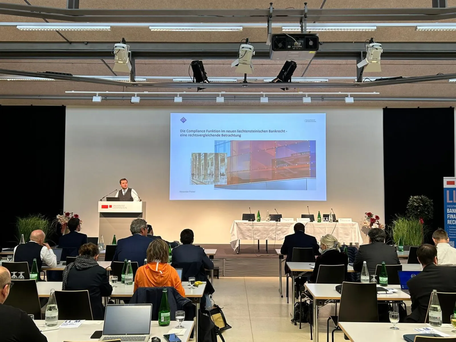 Ein Vortragender steht an einem Rednerpult vor einer Leinwand, auf der eine Präsentation gezeigt wird. Im Publikum sitzen mehrere Personen an Tischen mit Laptops. Die Veranstaltung findet in einem modernen Konferenzraum statt.