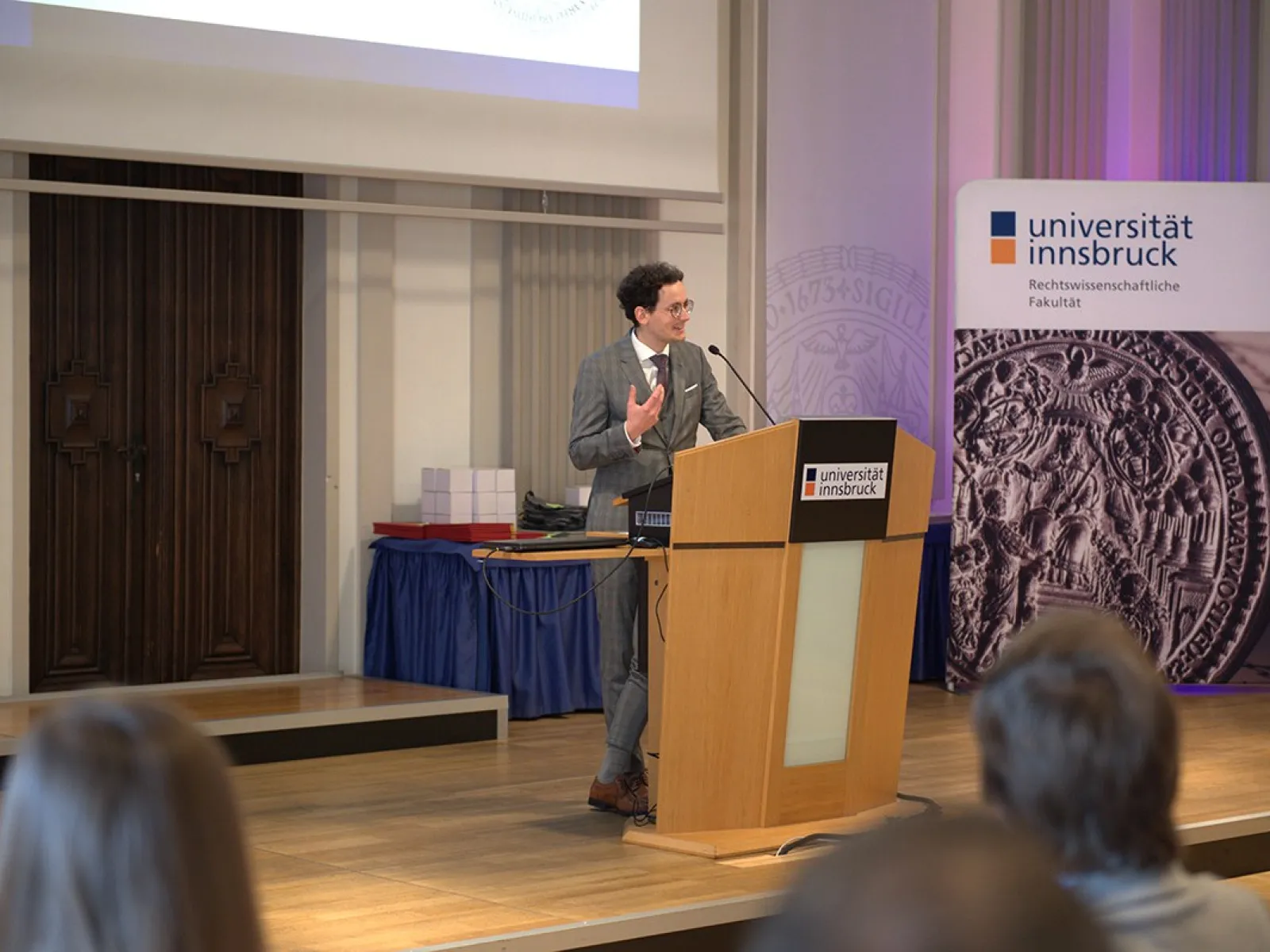 Ein Mann hält eine Präsentation an einem Rednerpult in einem Saal der Universität Innsbruck. Im Hintergrund sind ein Banner der Universität und ein Tisch mit Gegenständen zu sehen. Publikum sitzt im Vordergrund.