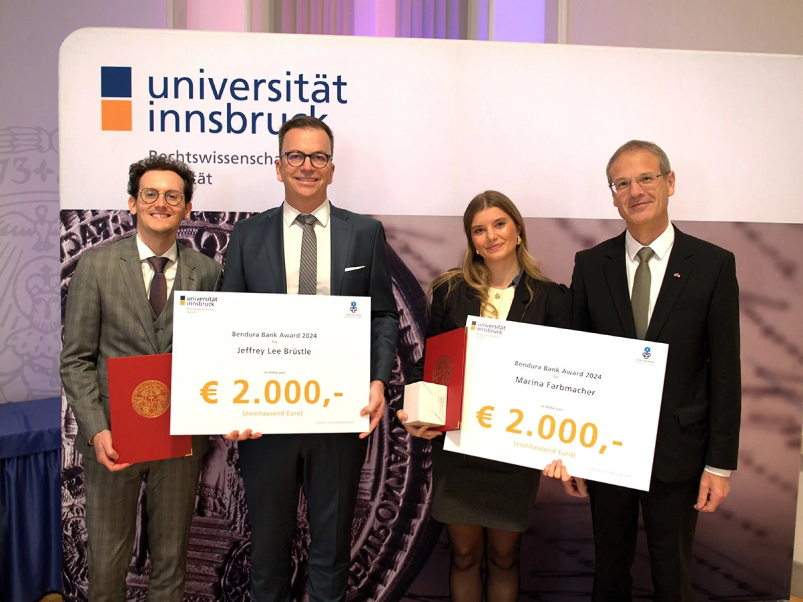 Vier Personen stehen nebeneinander und halten große Schecks mit der Aufschrift "Bendura Bank Award 2024" und einem Betrag von 2.000 Euro. Im Hintergrund ist ein Banner der Universität Innsbruck zu sehen. Die Personen sind formell gekleidet und lächeln in die Kamera.