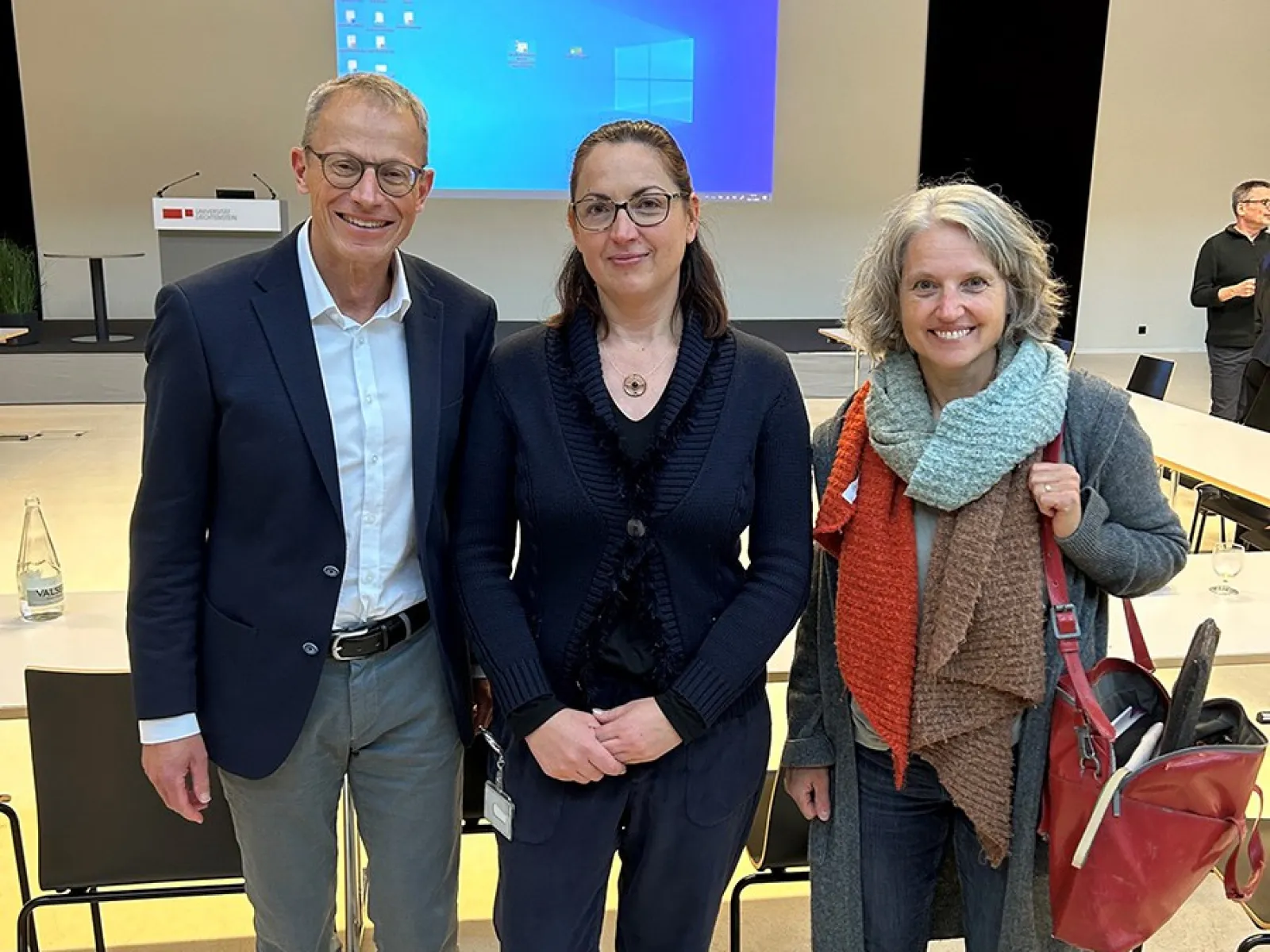 Drei Personen stehen lächelnd in einem Auditorium der Universität Liechtenstein. Im Hintergrund sind ein Rednerpult und eine Leinwand mit einer Präsentation zu sehen.