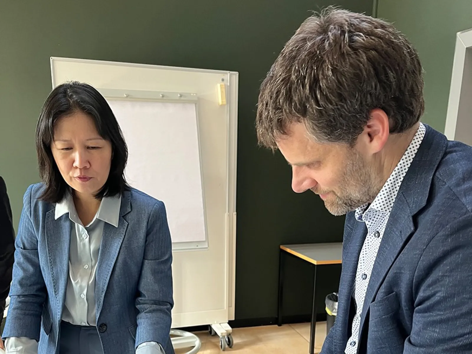 Zwei Personen in formeller Kleidung betrachten Bücher auf einem Tisch in einem Konferenzraum. Im Hintergrund ist ein Flipchart zu sehen.