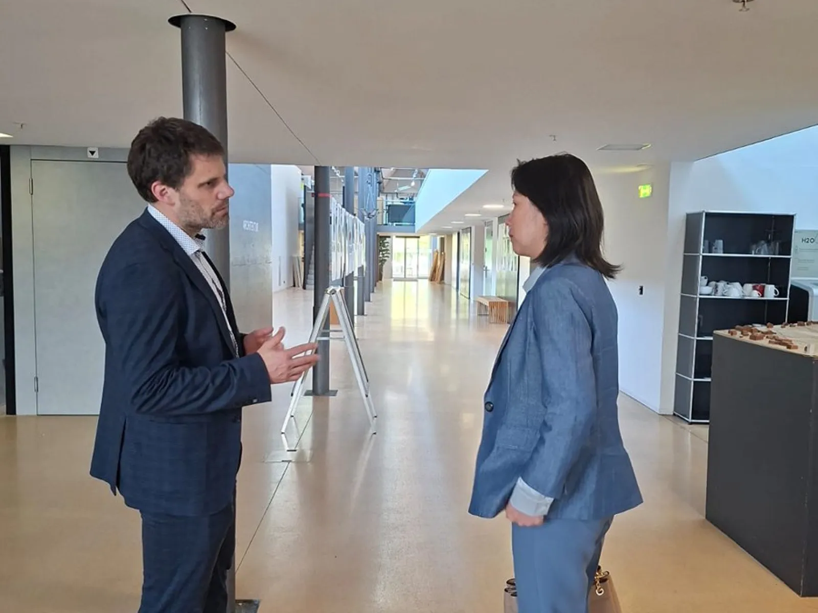 Zwei Personen in einem modernen Gebäude im Gespräch. Beide tragen Business-Kleidung. Im Hintergrund sind helle Flure und moderne Einrichtungselemente zu sehen.