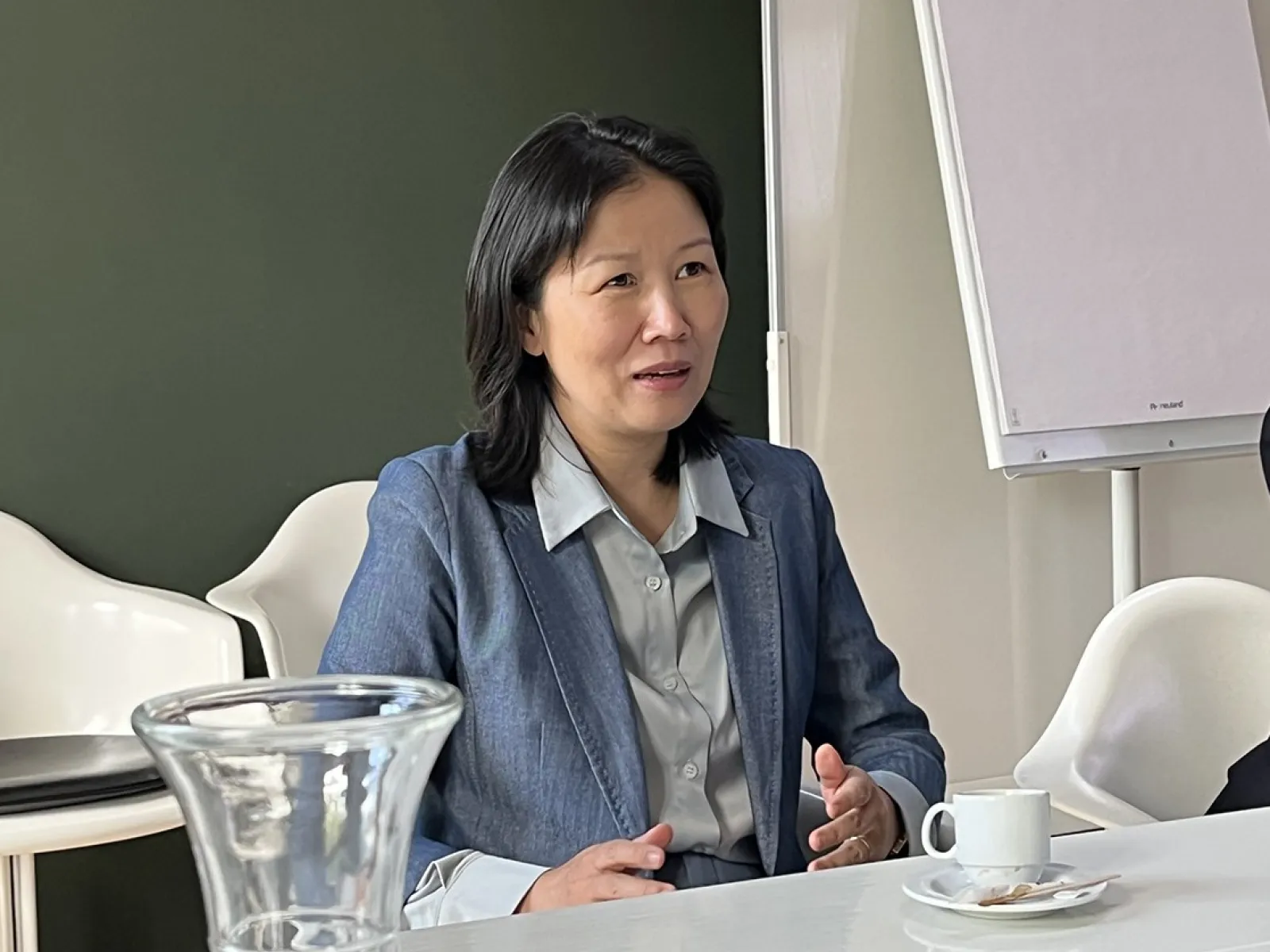 Eine Person in einem blauen Blazer sitzt an einem Konferenztisch mit einer Tasse Kaffee und einem Notizblock. Im Hintergrund ist ein Flipchart zu sehen.