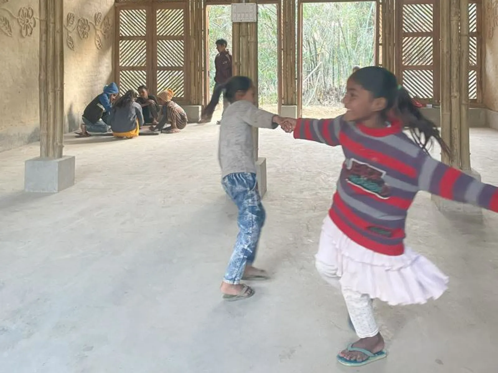 Kinder spielen und tanzen in einem neu gebauten Schulgebäude aus Bambus in Bihar, Indien. Im Hintergrund sitzen weitere Kinder auf dem Boden. Das Gebäude hat eine natürliche, lichtdurchflutete Atmosphäre.