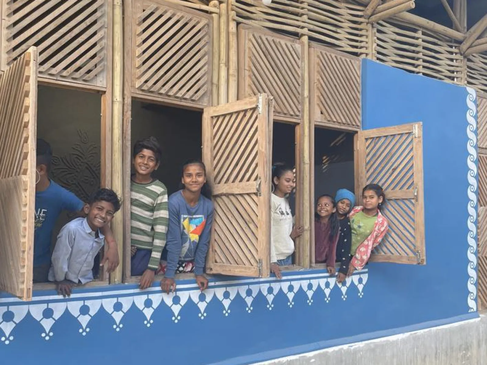 Kinder schauen lächelnd aus den Fenstern eines neu gebauten Schulgebäudes in Bihar, Indien. Die Fassade ist blau mit dekorativen weißen Mustern.