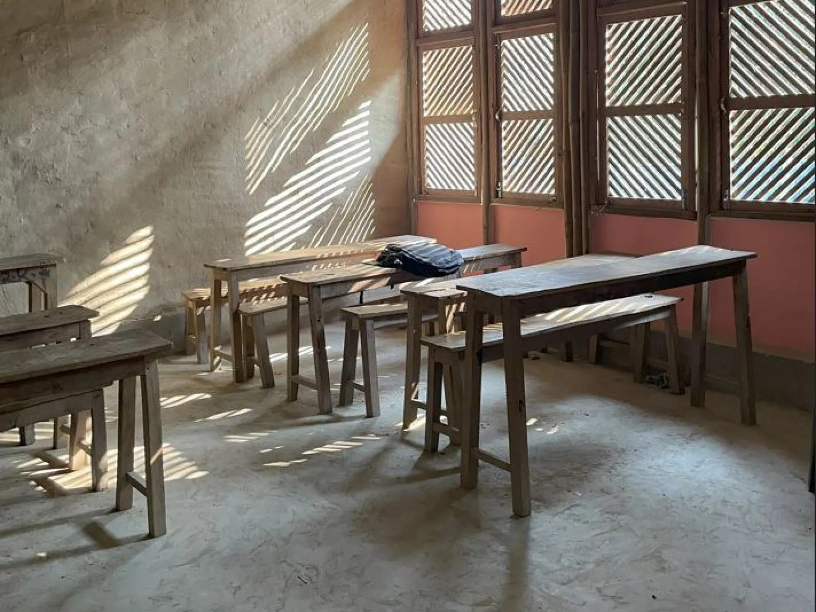 Ein einfacher Klassenraum mit Holztischen und -bänken, durchflutet von Sonnenlicht, das durch Fenster mit Holzlamellen fällt.