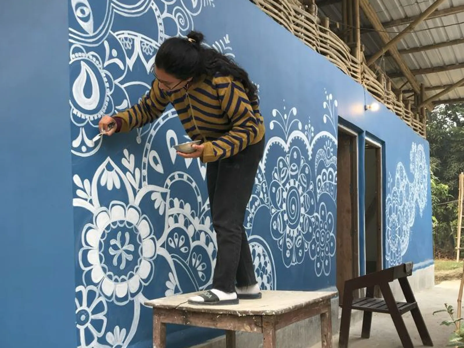 Eine Person bemalt die blaue Außenwand einer Schule in Indien mit weißen, floralen Mustern. Sie steht auf einem Holztisch, um die oberen Bereiche zu erreichen. Das Gebäude hat ein Wellblechdach und ist Teil eines Bildungsprojekts.