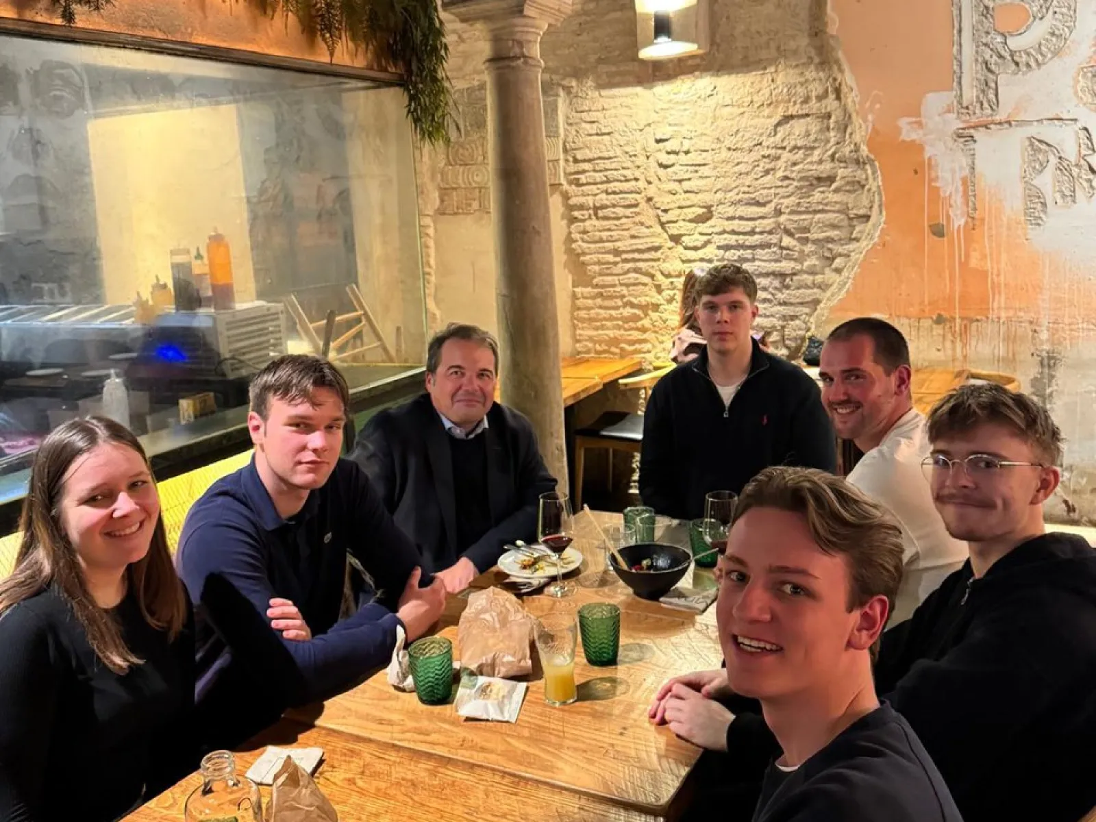 Eine Gruppe von sieben Personen sitzt an einem Tisch in einem Restaurant. Sie lächeln in die Kamera. Im Hintergrund sind eine gemauerte Wand und Pflanzen zu sehen.