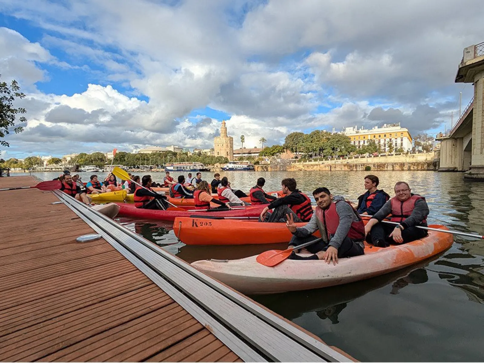 Studierende in Kanus auf einem Fluss in Sevilla, mit Schwimmwesten, vor einer malerischen Stadtlandschaft und bewölktem Himmel.