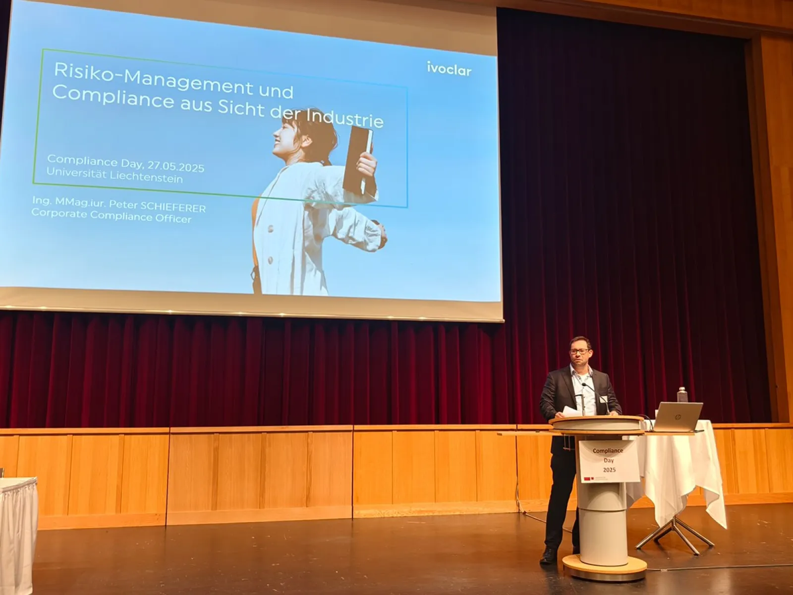 Ein Redner steht auf einer Bühne vor einem großen Bildschirm, der eine Präsentation mit dem Titel „Risiko-Management und Compliance aus Sicht der Industrie“ zeigt. Der Raum hat Holzwände und einen roten Vorhang. Ein Tisch mit Laptop und Mikrofon steht neben dem Redner.