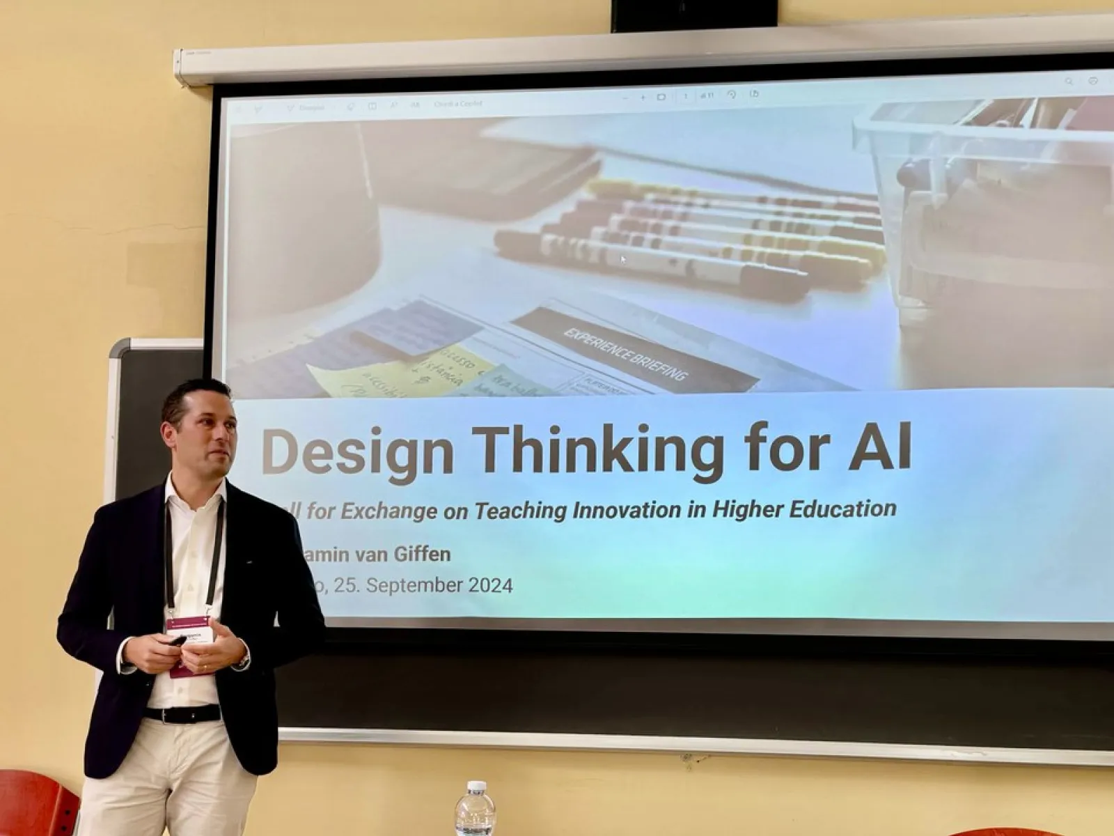 Ein Mann steht vor einer Leinwand, auf der eine Präsentation mit dem Titel "Design Thinking for AI" gezeigt wird. Der Raum wirkt wie ein Seminarraum, und es sind einige Stifte und Notizen auf der Leinwand zu sehen.