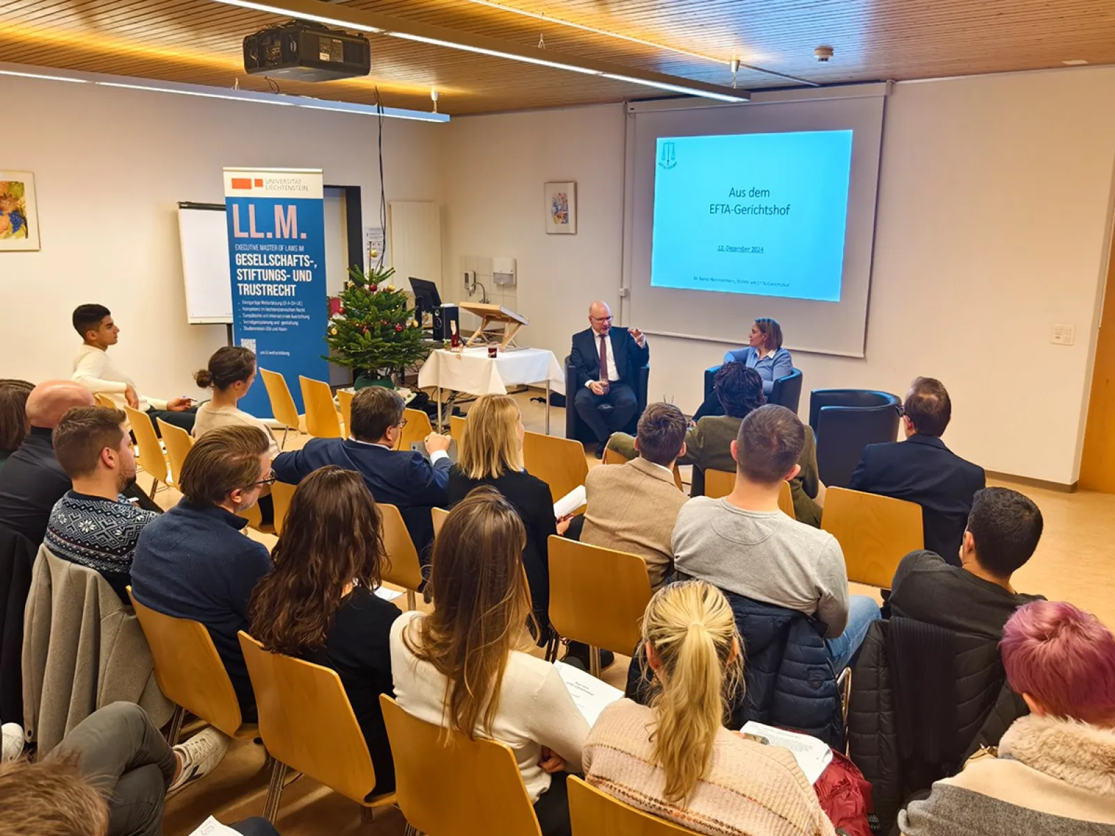 Ein Vortrag im Seminarraum der Universität Liechtenstein: Ein Redner und eine Rednerin sitzen vorne, während das Publikum aufmerksam zuhört. Im Hintergrund ist eine Präsentation mit dem Titel „Aus dem EFTA-Gerichtshof“ zu sehen. Ein Banner weist auf den LL.M.-Studiengang hin.