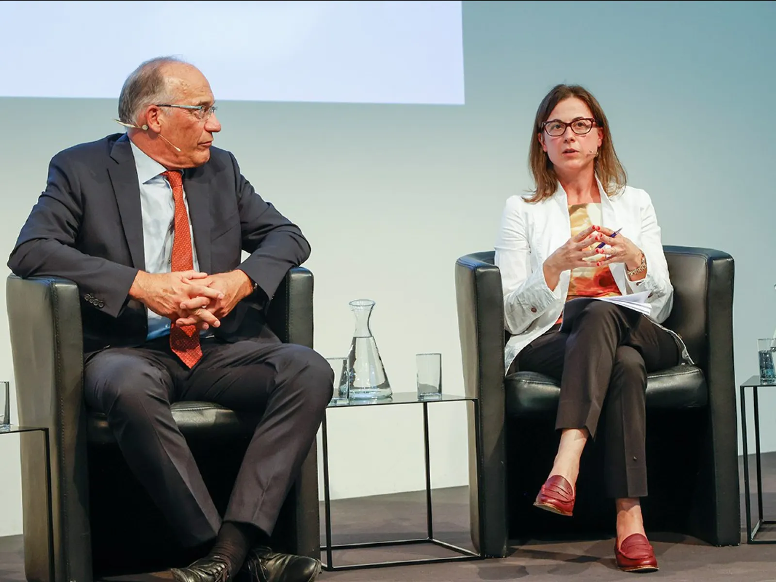 Zwei Personen sitzen auf einer Bühne in schwarzen Sesseln. Die Person links trägt einen dunklen Anzug und eine rote Krawatte, die Person rechts trägt eine weiße Jacke und rote Schuhe. Zwischen ihnen stehen Wasserkaraffen und Gläser.