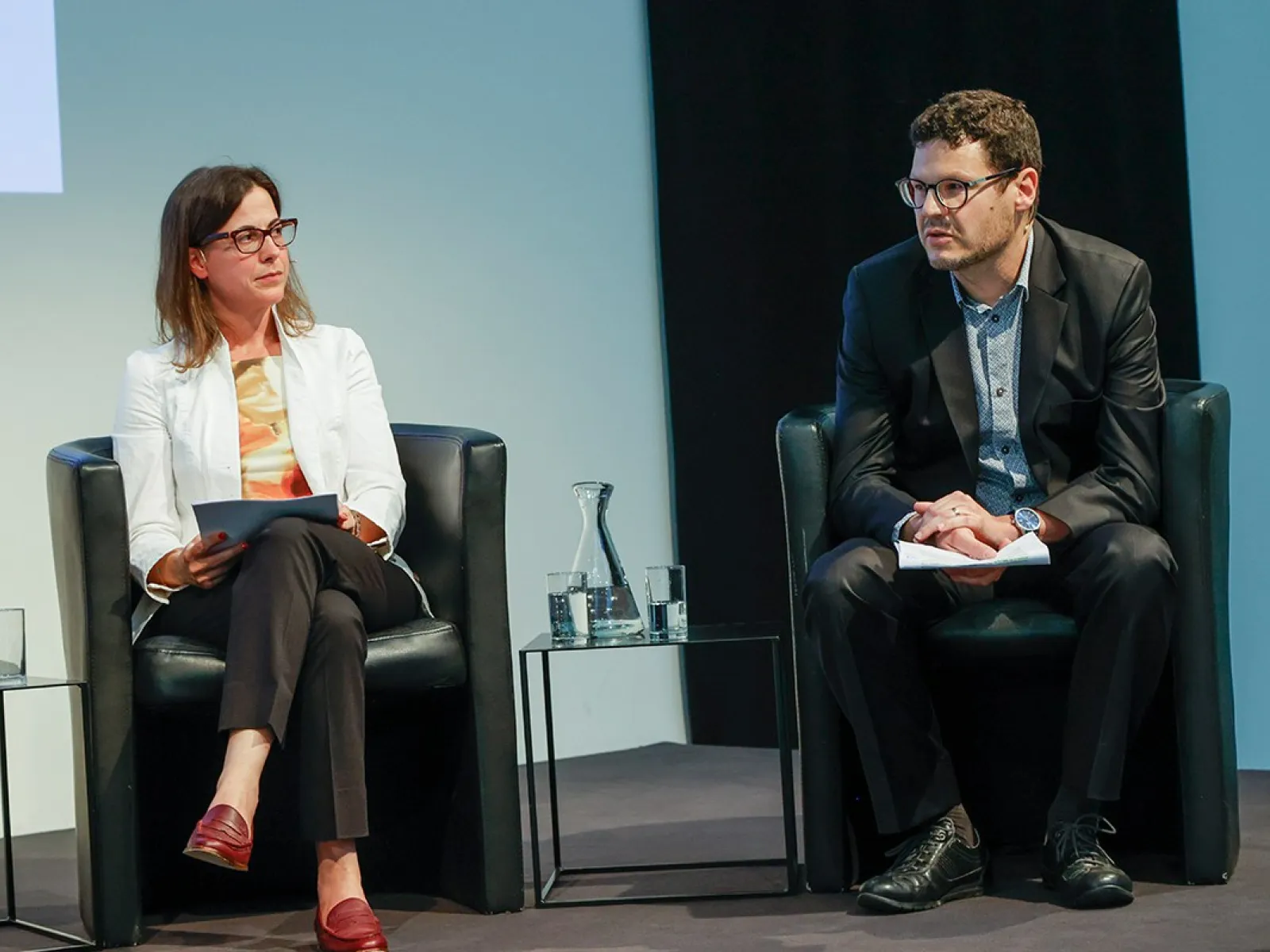 Zwei Personen sitzen auf einer Bühne in schwarzen Sesseln. Die Frau links trägt eine weiße Jacke und hält Papiere, der Mann rechts trägt einen dunklen Anzug. Zwischen ihnen steht ein Wasserglas auf einem kleinen Tisch.