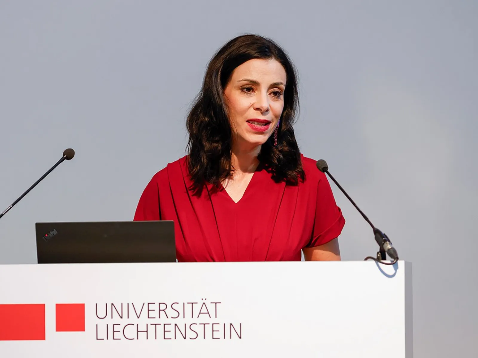 Eine Frau in rotem Kleid spricht an einem Rednerpult mit dem Logo der Universität Liechtenstein. Sie hält eine Rede vor einem Mikrofon.