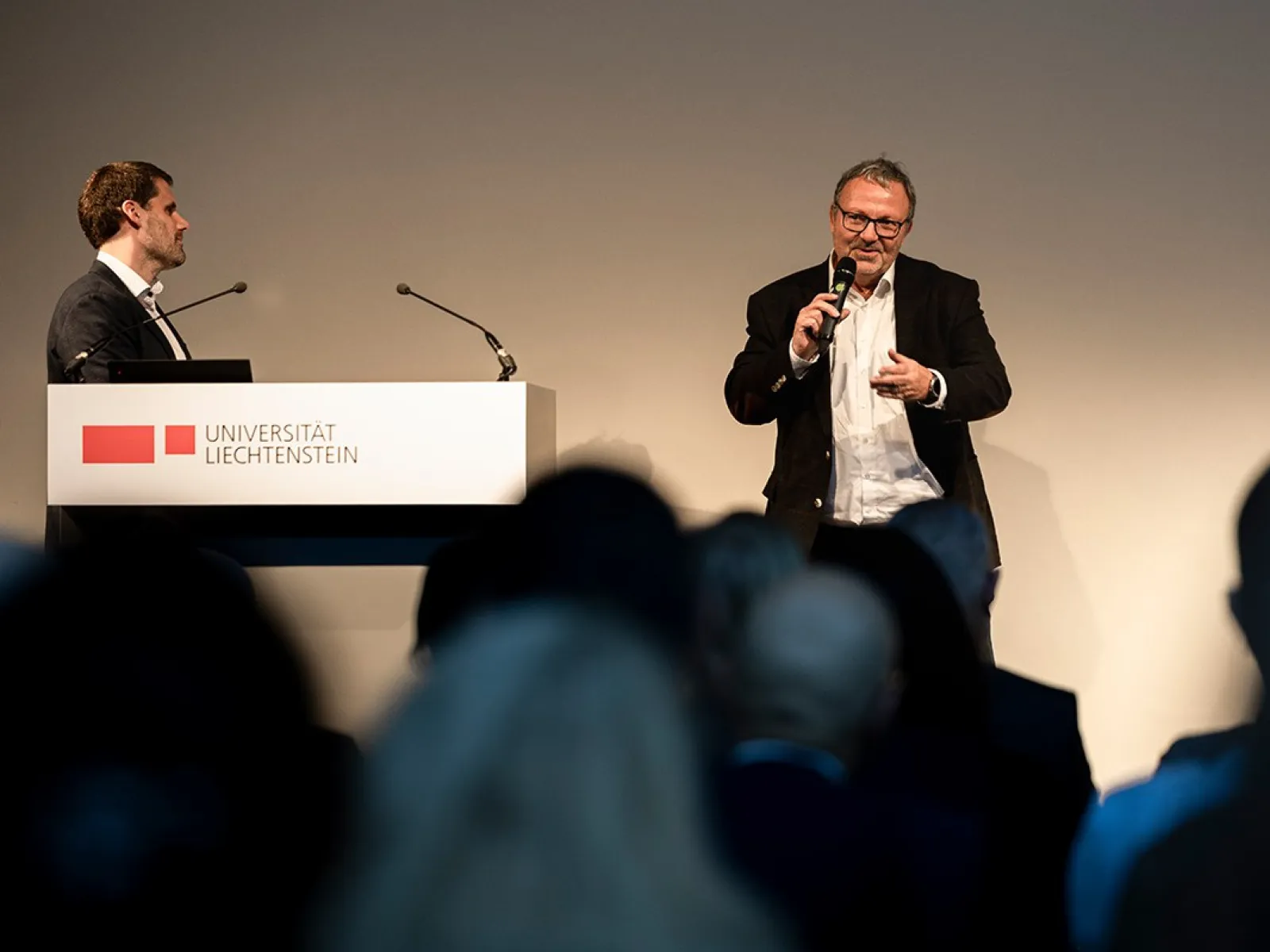 Zwei Männer stehen auf einer Bühne bei einer Veranstaltung an der Universität Liechtenstein. Einer spricht in ein Mikrofon, während der andere neben einem Rednerpult mit dem Logo der Universität steht. Im Vordergrund sind unscharf die Köpfe des Publikums zu sehen.