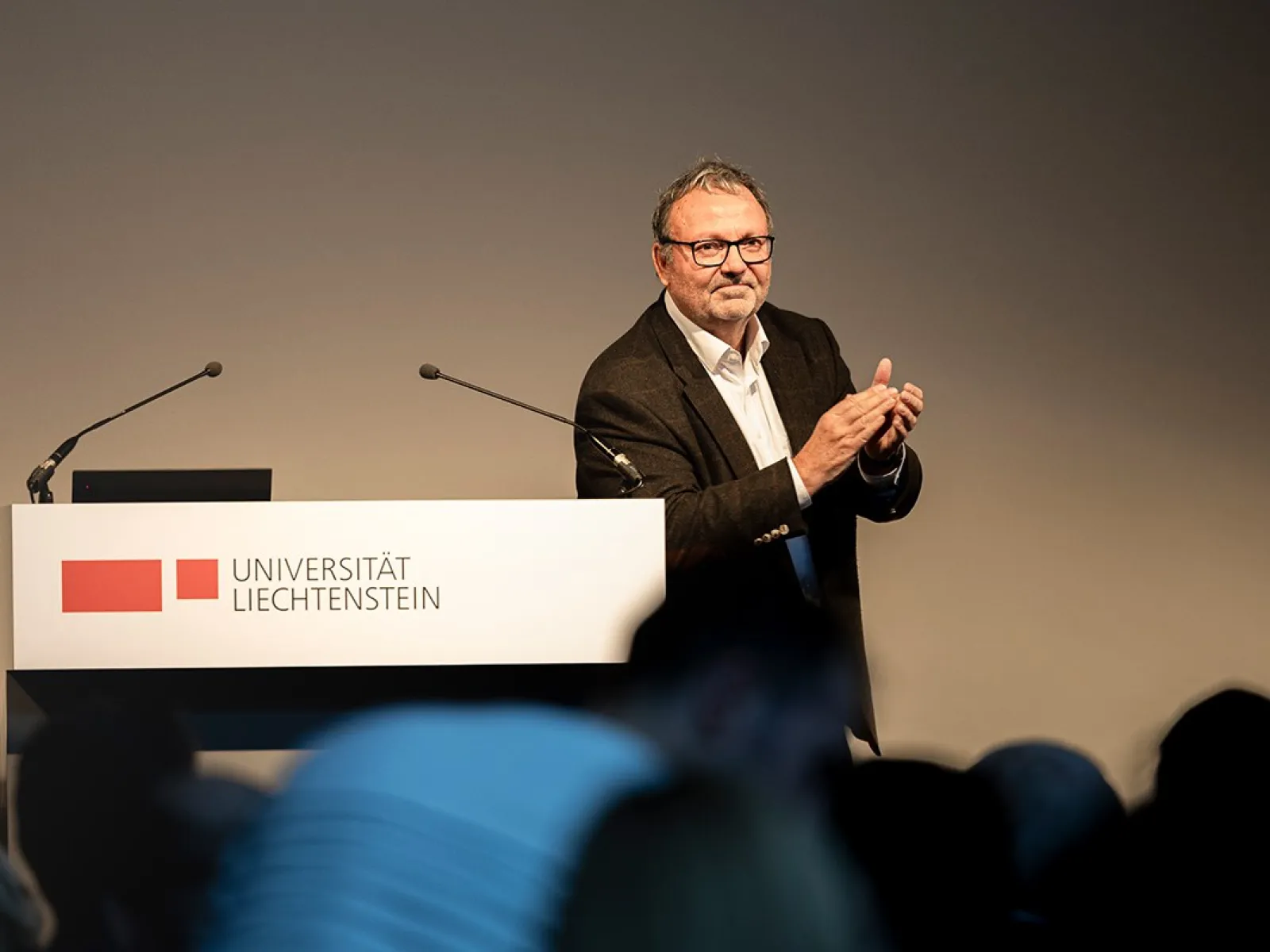 Ein Mann steht an einem Rednerpult der Universität Liechtenstein und applaudiert. Im Hintergrund sind Mikrofone sichtbar.