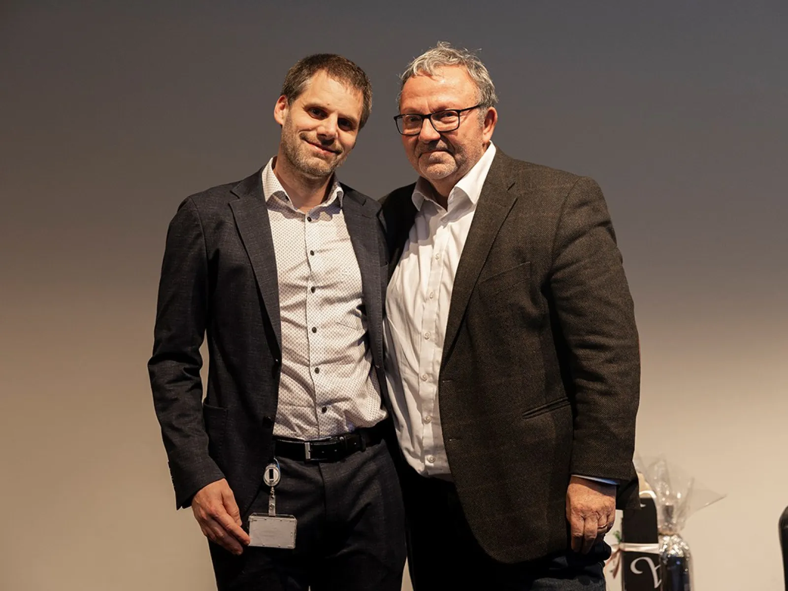 Zwei Männer stehen nebeneinander und lächeln in die Kamera. Sie tragen Anzüge und befinden sich in einem Innenraum mit neutralem Hintergrund.